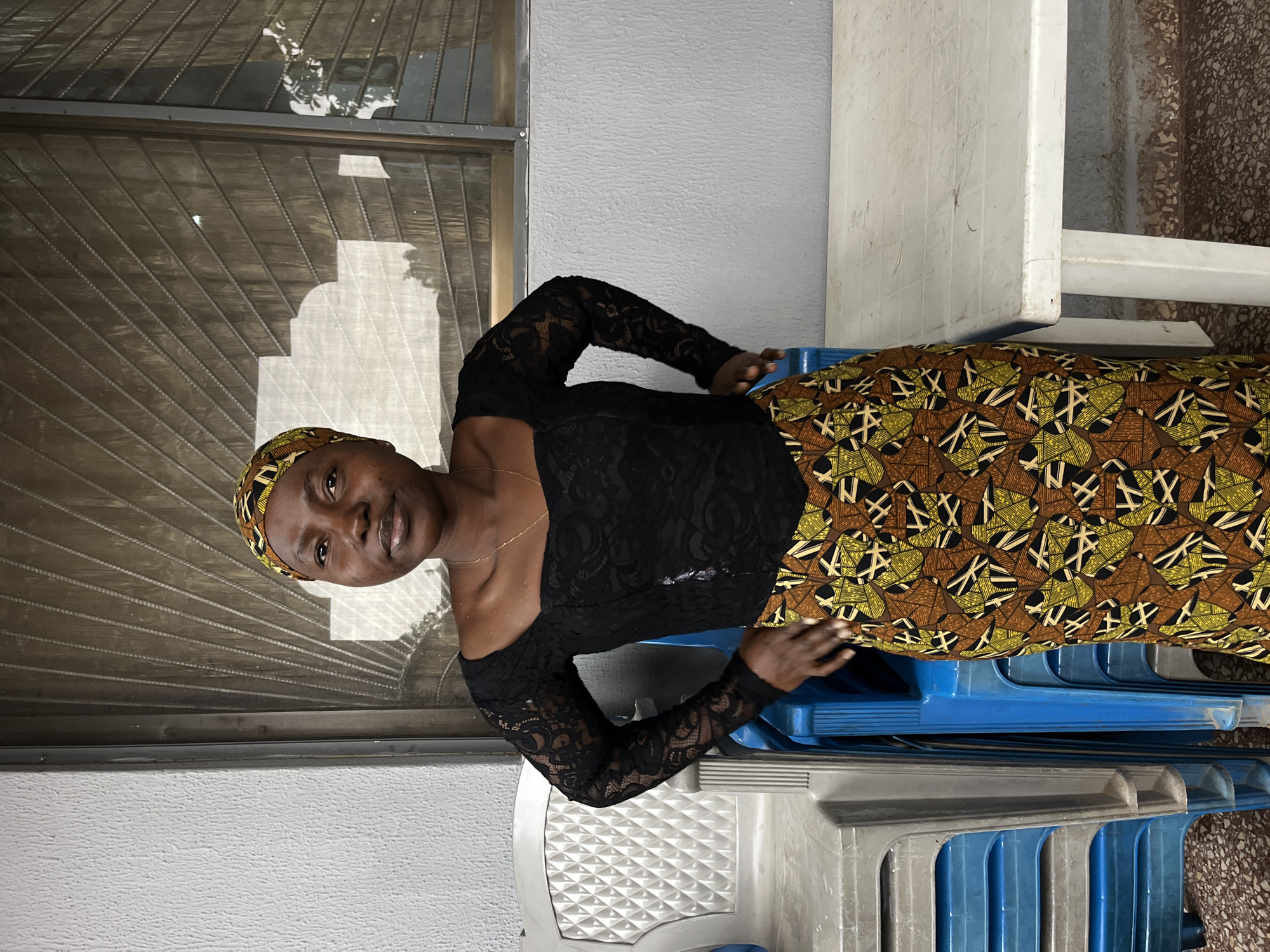 A medium shot captures a Black woman from the chest up, standing with her hands on her hips in front of a window. She wears a black lace top with long sleeves and a brown and yellow patterned skirt. A colorful headscarf adorns her hair. The background features a window with a metal grate, and to the left, a stack of blue and white plastic chairs is visible, along with a white plastic table on the right. The setting appears to be an outdoor or semi-outdoor area.