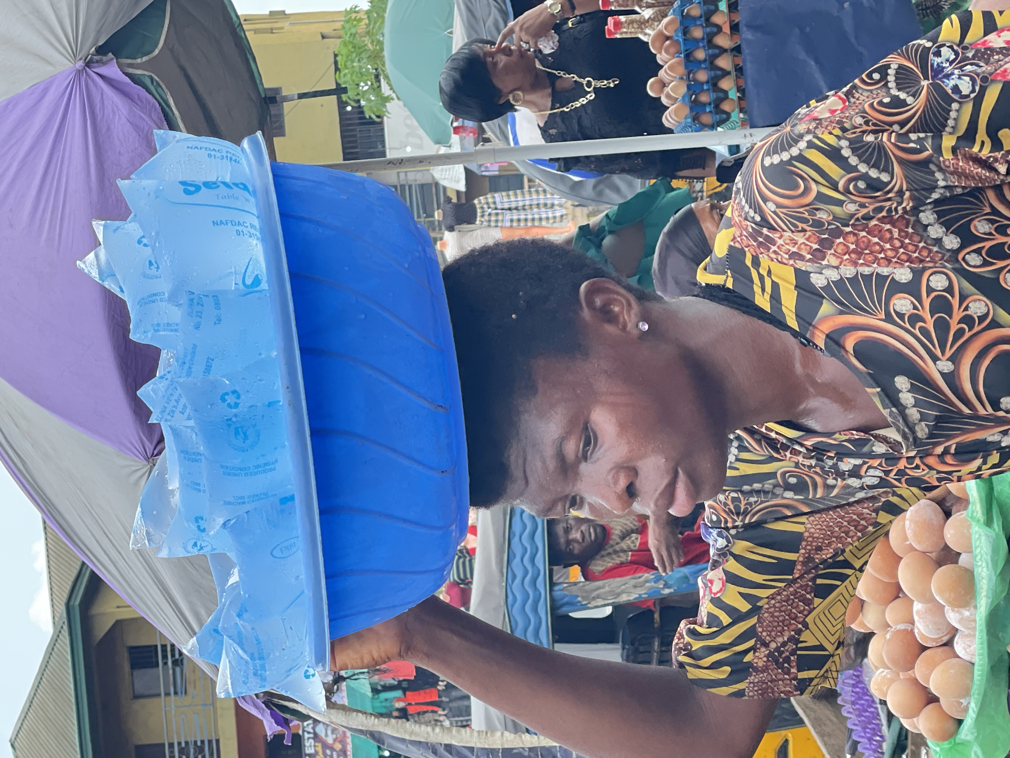 The image depicts a bustling outdoor market scene in the city of Unknown, Unknown, likely under partial shade from market umbrellas. The central figure is a young person with dark skin and short, dark, kinky hair, seen in profile, facing right. They are carrying a vibrant blue plastic basin on their head, which is filled with numerous clear plastic sachets of cold drinking water. Condensation and slight frosting are visible on the sachets, some bearing text such as "NARDAC PR," "Table Water," and a recycling symbol. The person is visibly sweating, with moisture on their forehead, cheek, and neck. They wear a short-sleeved top with an intricate, multi-colored pattern in yellow, orange, brown, and black. A small, light-colored stud earring is present in their left earlobe, and their left arm is raised, with the hand lightly touching the basin for support.
In the lower-left foreground, a green plastic bag contains a mound of brown eggs, some exhibiting a white dusty residue. To the right of the central figure, a woman with dark skin, short dark hair, and prominent gold hoop earrings is visible. She wears a black lace-patterned top and a gold-colored necklace with a large pendant. Her right hand is near her mouth or nose, and she is looking towards the right. Behind her, a blue plastic tray holds neatly arranged brown eggs.
In the background, various elements of the market are visible. Overhead, parts of large market umbrellas in purple, grey, and green provide shade. To the left, a multi-story building with a brown/green roof and a balcony is present, along with a partial sign displaying "ESTA." Behind the central figure, another individual, a man in a red shirt with a patterned scarf around his neck, is partially visible, looking towards the viewer. Further in the background, a yellow building with windows and a metal gate is discernible. Mannequins displaying a light-colored top and plaid trousers are visible on a stall in the mid-ground, suggesting a clothing vendor. The overall environment suggests a vibrant commercial area.