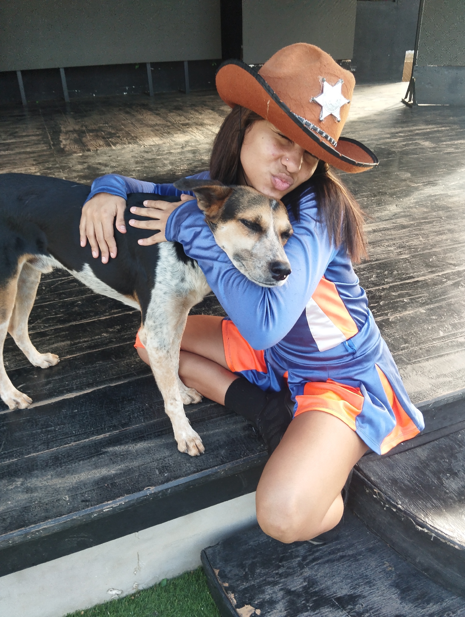 A person wearing a brown cowboy hat and a blue and orange outfit is sitting on a wooden surface with a dog. The person is embracing the dog, who appears to be resting its head against the person's chest. The person is making a kissing face. The wooden surface appears to be a stage or platform. There is a dark, undefined backdrop behind them and a patch of green grass visible at the bottom edge of the frame. The location is Unknown, Unknown.
