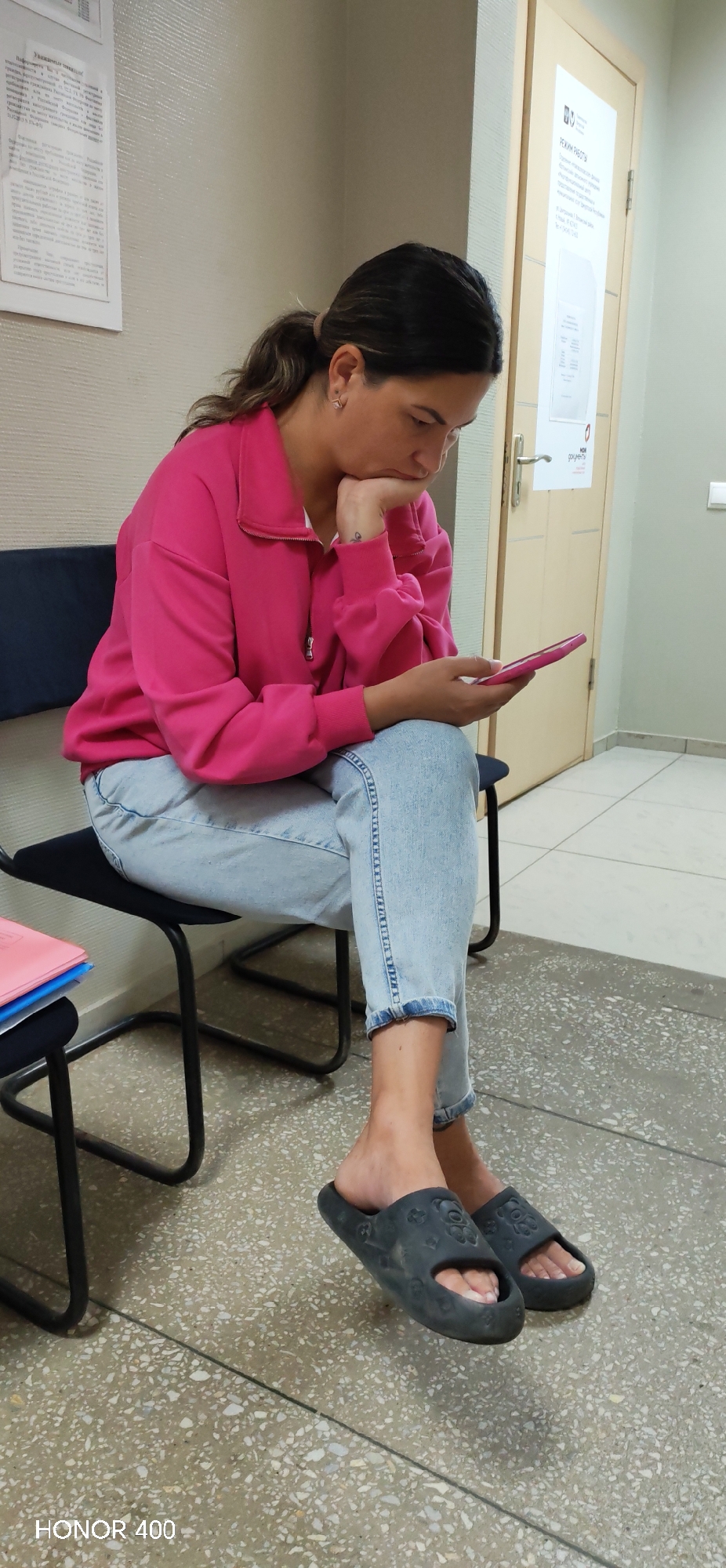 A woman is seated on a dark chair, looking at her pink smartphone. She is wearing a bright pink zip-up sweatshirt and light blue jeans that are cuffed at the ankles. Her feet are adorned with dark gray slides, each decorated with a subtle bear-shaped pattern. Her right hand rests on her chin, and her left hand holds the phone. A small tattoo is visible on her left wrist. To her left, on an adjacent chair, are stacked folders, one pink and one blue. The environment appears to be an indoor waiting area, indicated by the beige, textured wall behind her and a light-colored door with a sign on it to her right. The floor is a mottled gray terrazzo. The overall lighting suggests an indoor setting during the daytime. The image includes a watermark in the bottom left corner: "HONOR 400". The location is identified as Noviy, Russia.
