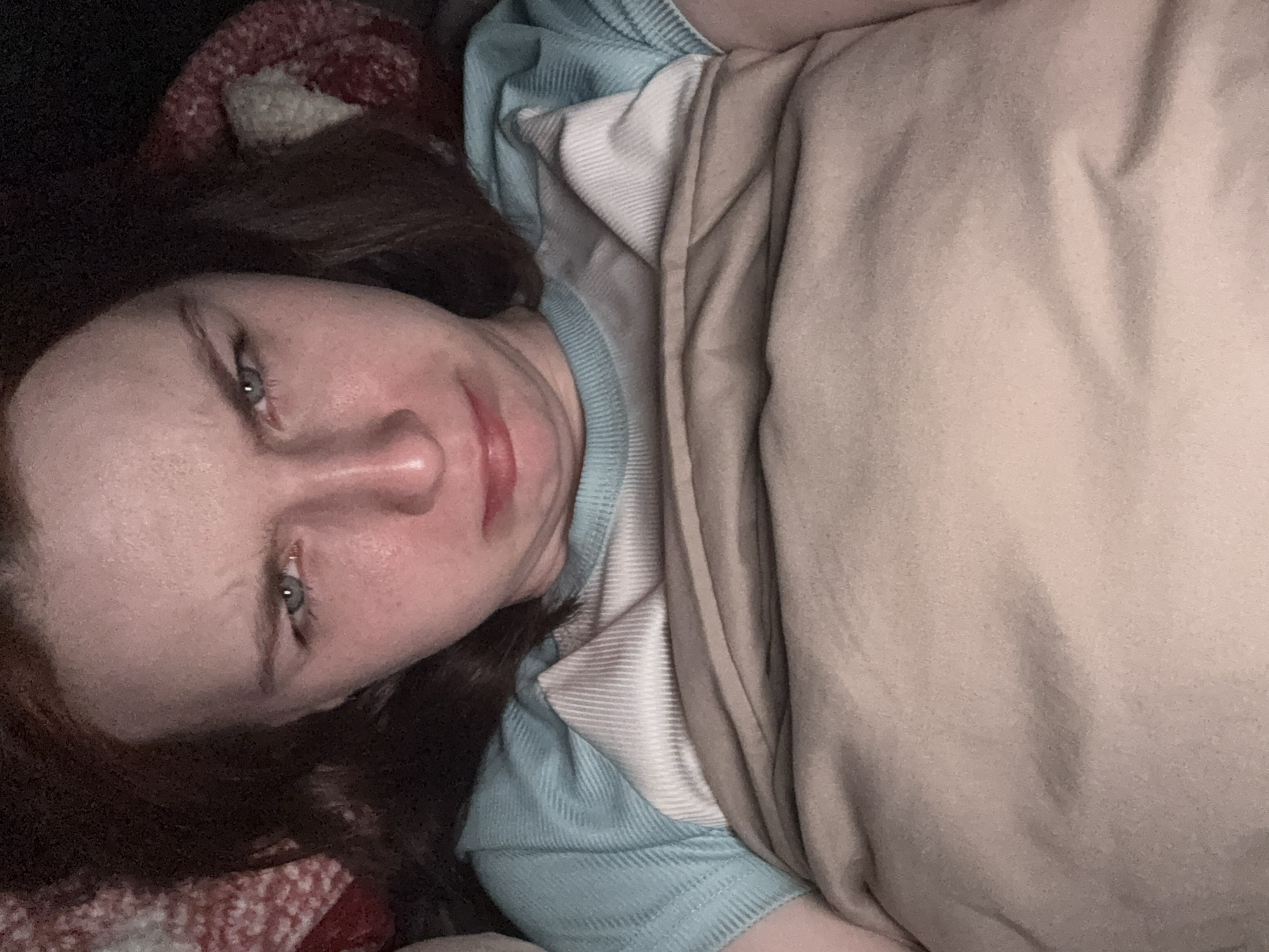 The image, taken in Lednevo, Russia, shows an adult female lying down indoors. Her head is slightly tilted to the left, and she is looking directly at the camera with a neutral expression that hints at slight weariness or a subtle frown. She has reddish-brown hair and light-colored eyes.
She is wearing a light blue garment, possibly a shirt or sweater, featuring white ribbed detailing around the neckline and shoulders, suggesting a layered appearance or a specific fabric design. A plain light brown or beige blanket covers her lower body, from her upper torso downwards. In the upper left background, behind her head, a reddish-brown textured blanket or textile with a faint pattern is visible.
The lighting in the scene is dim, suggesting it is either evening or that the room is dimly lit. No visible text is present in the image. The individual appears to be in a relaxed or resting position, possibly in bed or on a couch.