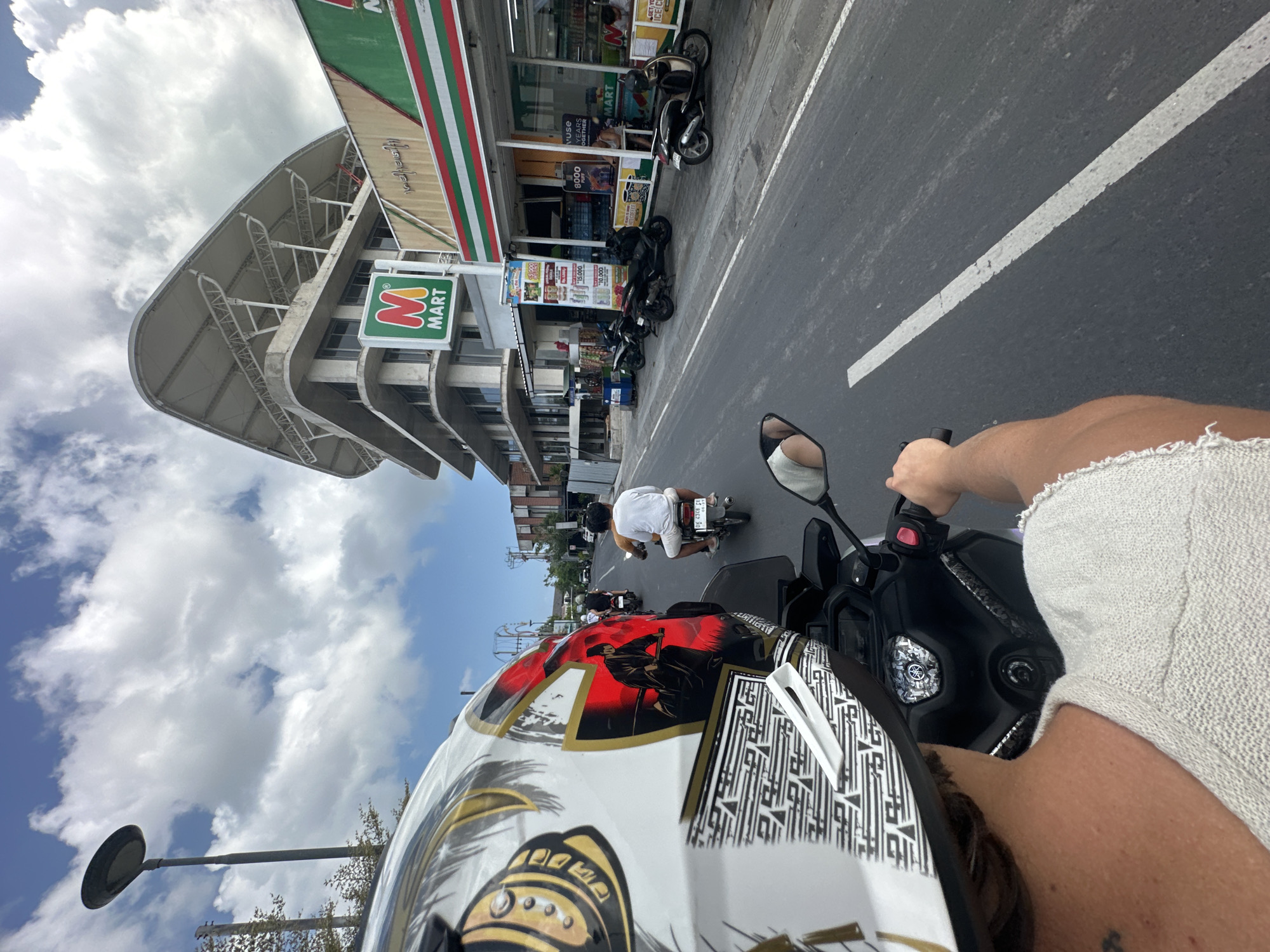 The image depicts a perspective from a person riding on a scooter, looking forward. In the foreground, a portion of the rider's helmet and shoulder is visible, showcasing a distinctive design with samurai imagery. The rider's arm is extended, holding the scooter's handlebar. The scooter itself is black with some reflective elements.
The scene is set on a paved road in North Kuta, Indonesia, characterized by a white lane marker. Several other scooters are visible on the road, with riders traveling in the same direction. To the right of the road, there is a row of buildings. The most prominent structure is a multi-story building with a distinctive, curved white roof. Below this, a commercial establishment with a green and white striped awning is visible, identified by a prominent "M MART" sign. This building appears to be a convenience store.
Further down the road, other buildings and trees are visible under a bright, partly cloudy sky. The overall atmosphere suggests a sunny day in an urban or semi-urban environment. The presence of multiple scooters and convenience stores indicates a busy, commercial area.