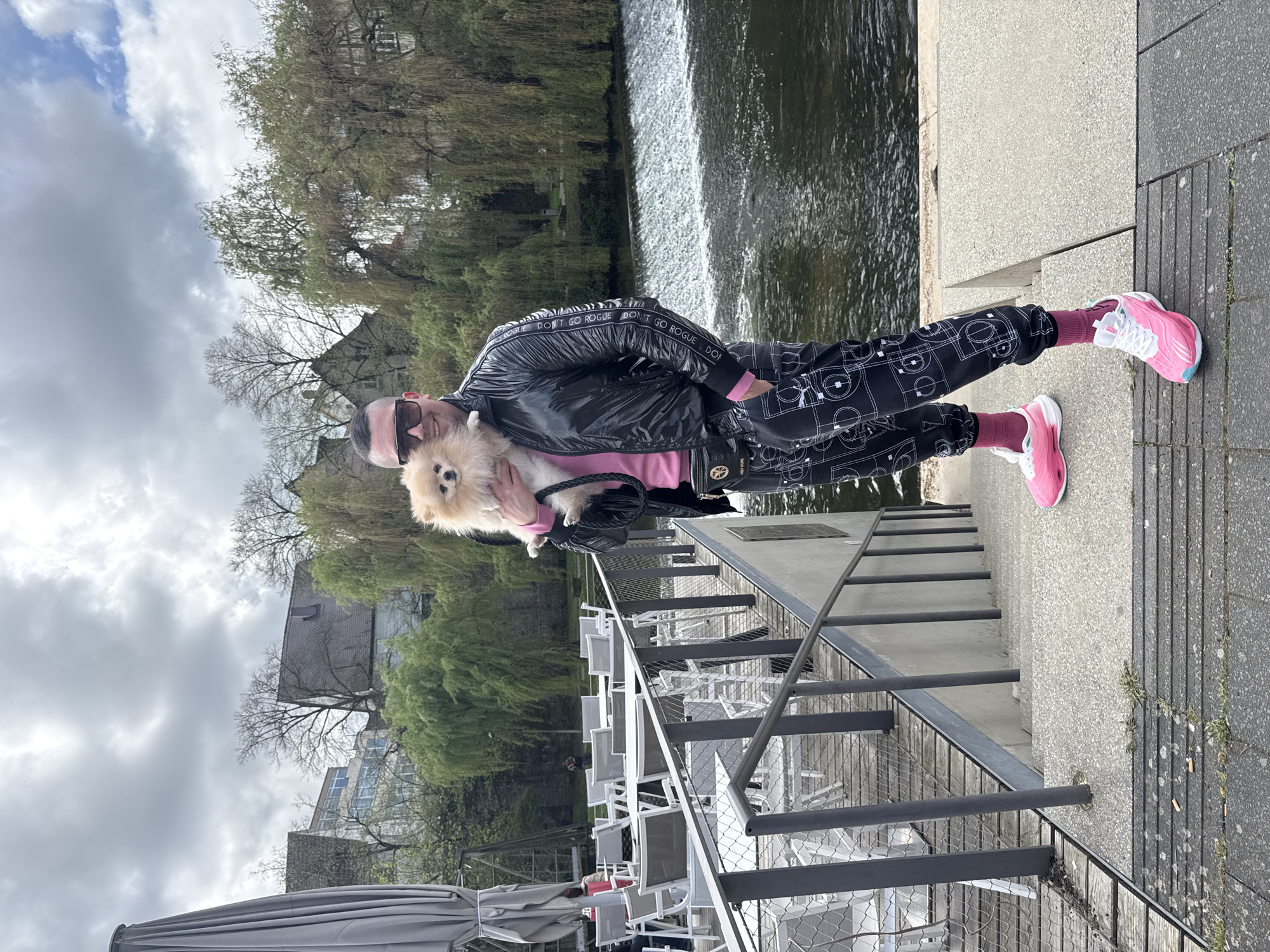 The image shows a person standing on a waterfront promenade in Backnang, Germany. The person is holding a small, light-brown Pomeranian dog. The person is wearing a black shiny jacket with white text that reads “DON’T GO ROGUE” repeatedly along the sleeves, black pants with white geometric patterns, pink socks, and pink and white sneakers. They are also wearing sunglasses.
Behind the person is a body of water, likely the Murr River, with a small waterfall or weir visible in the background. Trees line the opposite bank of the river. To the side of the person is a patio or deck area with metal railings, a partially visible umbrella, and numerous white chairs and tables, indicating a restaurant or cafe. The sky is overcast with a mix of clouds. Buildings are visible in the distance on the opposite bank.