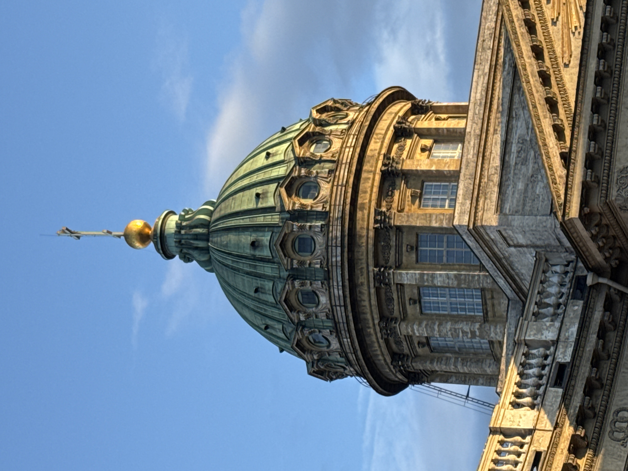 This is a close-up, low-angle, exterior, daylight, vertical, full-color, photographic image of the dome and upper section of the Kazan Cathedral in Saint Petersburg, Russia. The sky is a clear, bright blue with a few wispy clouds. The dome is large and green, with a golden orb and cross at its apex. The dome is supported by a circular, stone drum that features ornate architectural details, including windows and columns. Below the drum, a section of the cathedral's facade is visible, characterized by stone construction, windows, and decorative railings. The building appears to be bathed in warm sunlight, casting shadows and highlighting the architectural textures.