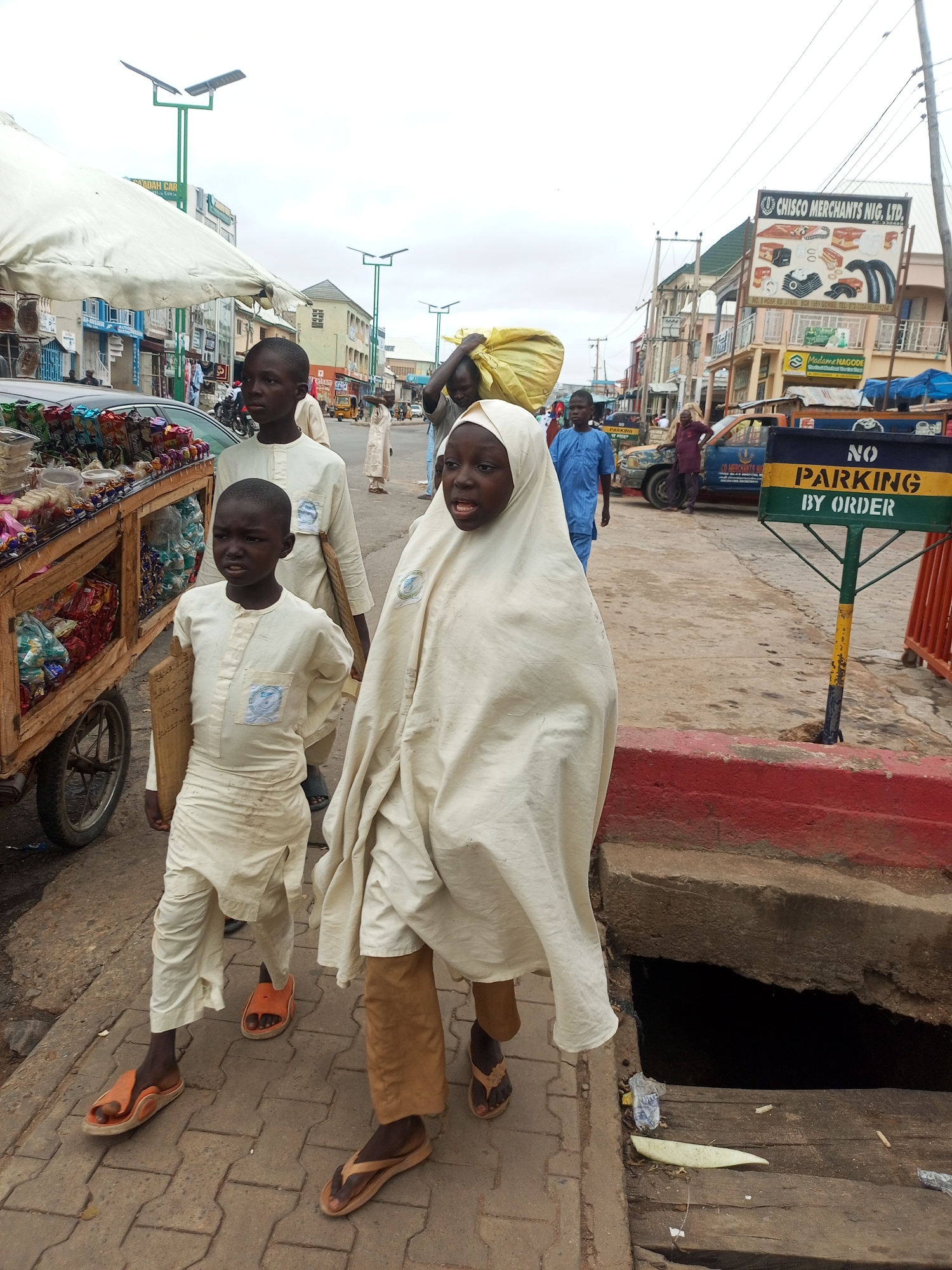 In Arawa, Nigeria, a street scene unfolds with a group of children walking along a sidewalk. The foreground is dominated by a young girl draped in a cream-colored hijab and robe, her expression one of focused determination as she walks forward. To her left, a boy in a similar cream-colored outfit, holding a wooden board, glances sideways with a serious expression. Behind him, another boy in a light blue outfit walks further down the street. A market stall laden with packaged goods, including colorful snacks and other items, is visible to the far left, partially obscured by a white awning. In the background, various buildings line the street, some with shops and signage, including a prominent sign for "CHISCO MERCHANTS NIG. LTD." A "NO PARKING BY ORDER" sign with green and yellow bands stands near the right edge of the frame. The overall environment suggests a busy urban marketplace with typical street elements like paved walkways, vehicles, and overhead wires. The sky is overcast.