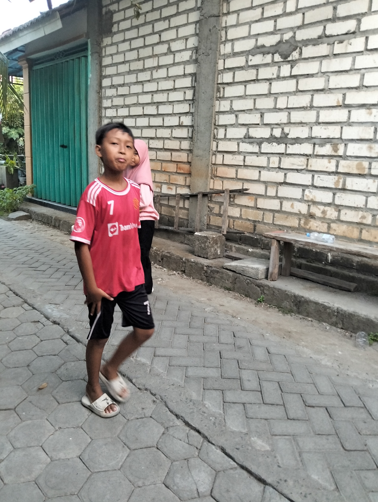 In Paciran, Indonesia, a young boy wearing a red t-shirt with white stripes on the sleeves and the number "7" prominently displayed, along with black shorts, walks on a paved walkway. His shirt has a logo and the word "Bamins" visible. He is wearing white slide sandals. Behind him, a person wearing a pink hijab and a light-colored shirt is partially visible. They are standing in front of a white brick wall. To the left, a green metal door is visible. The walkway is made of hexagonal concrete pavers. A wooden bench with a plastic bottle on it is situated against the brick wall. The scene appears to be an outdoor, possibly residential or public space, with evidence of construction or repair work in the background.