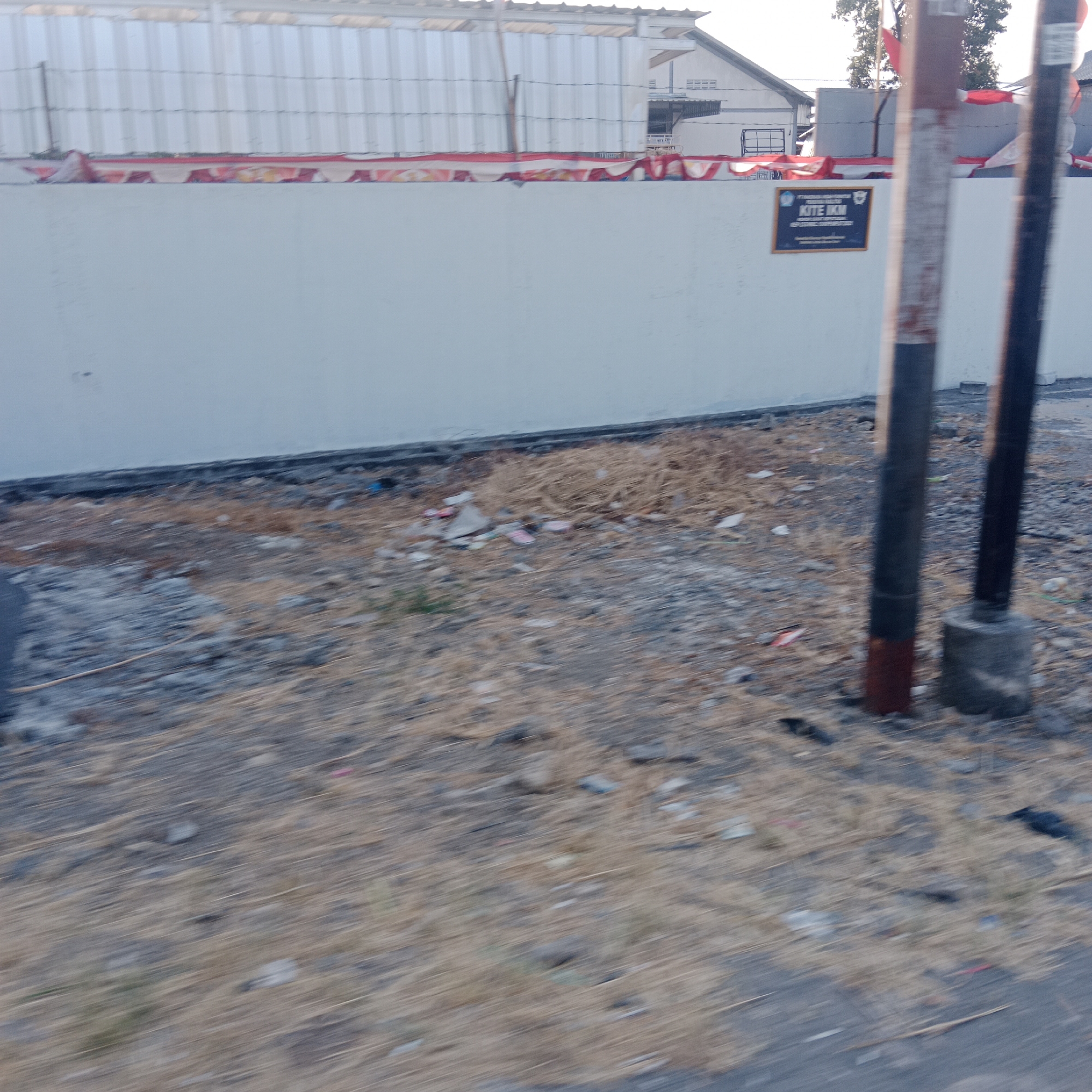 The image shows a partially blurred view of a construction site or newly built area. In the foreground, dry grass and some debris are scattered on the ground. Two vertical poles, one red and white and the other darker, are prominent in the middle ground, with one anchored in a concrete base. A light-colored wall runs across the scene, decorated with a red and white banner draped along its top edge. On the wall, a dark rectangular sign with a yellow border displays text, appearing to read "KITE IKM" and further information below. In the background, a corrugated metal structure is visible, likely part of a building, with a white building with visible windows and louvers behind it. The overall impression is of an unfinished or recently completed outdoor space. The image is taken from a slightly elevated perspective, possibly from a moving vehicle, contributing to the blurriness. The location is stated to be in Lobangdolok, Indonesia.