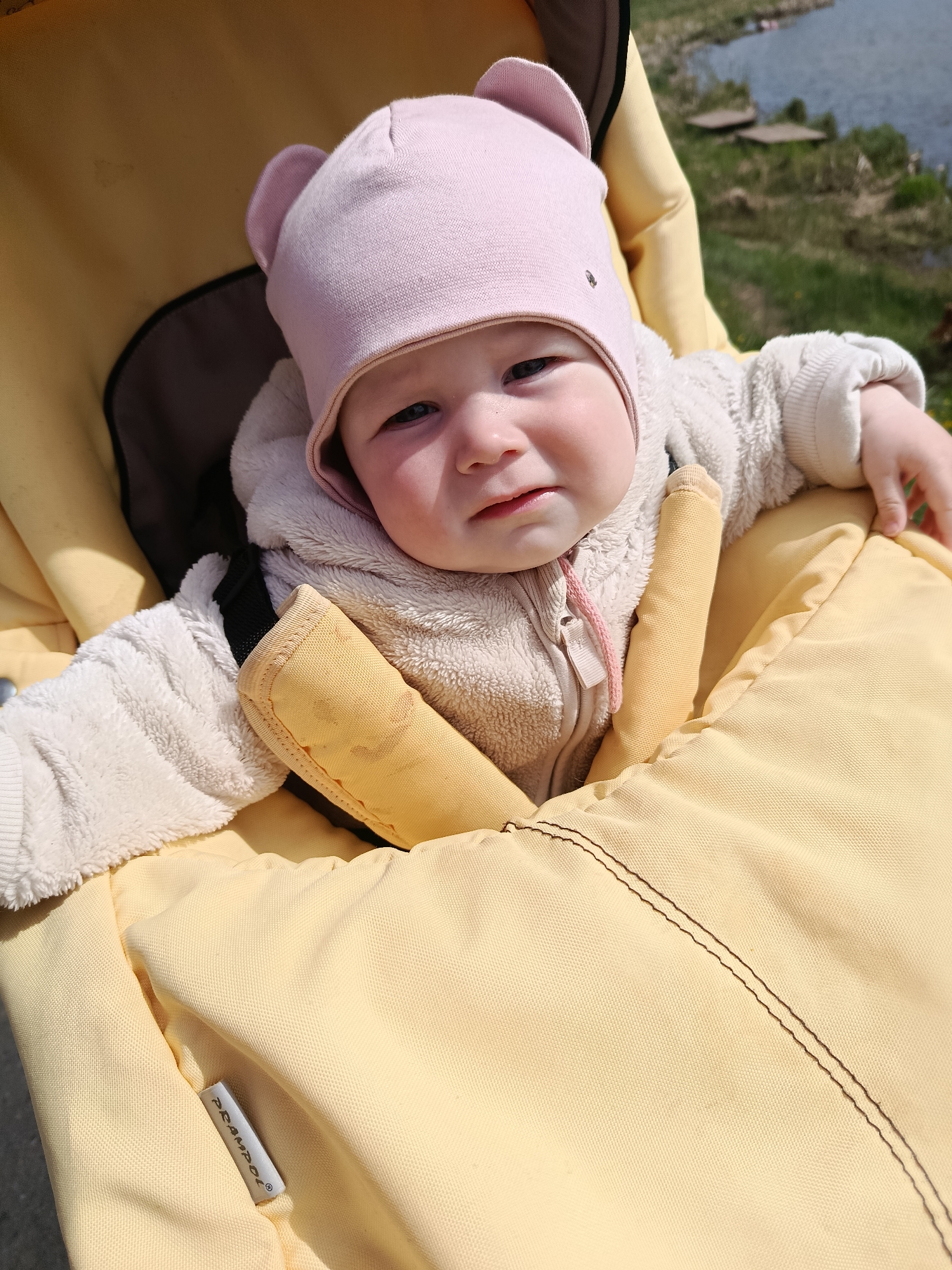 A baby wearing a pink hat and a cream-colored jacket is secured in a yellow stroller. The stroller has yellow harness pads and a yellow cover. In the background, out of focus, is a body of water, likely a lake or river, with grassy vegetation on the bank.