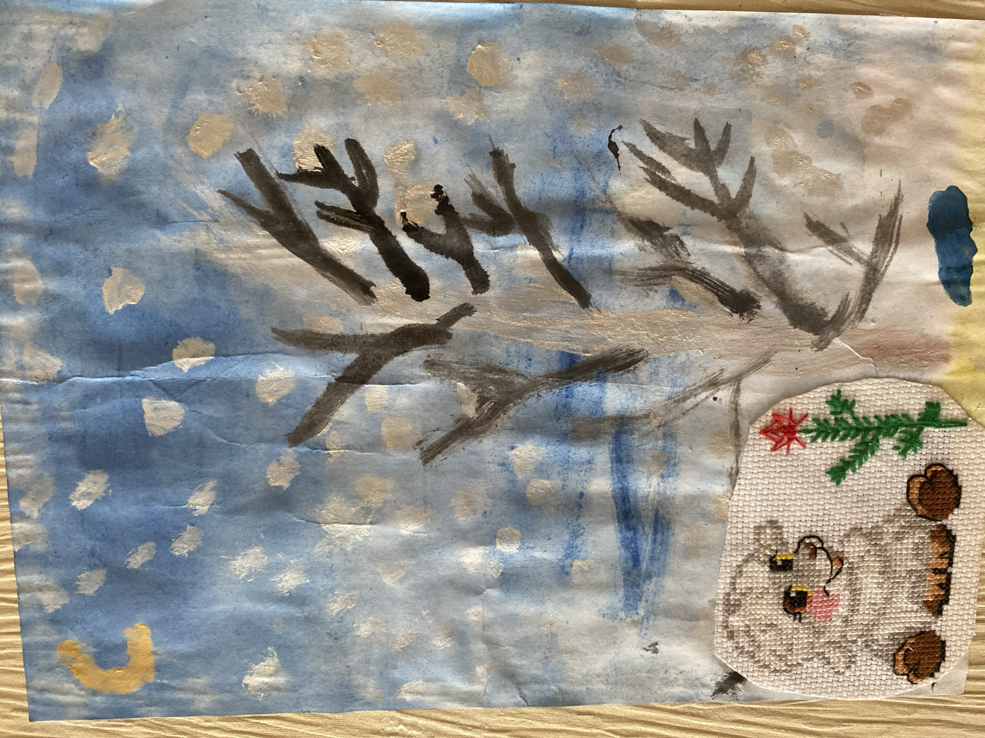 This is a close-up, top-down view of a crumpled piece of paper with a watercolor painting on it. The painting depicts a winter scene with a bare tree in the center. The sky is a mottled blue with white and pale yellow splotches that could represent clouds or snow. A yellow crescent moon is in the upper left corner. The foreground is a pale yellow and blueish-gray area. In the lower left corner of the painting is a separate, oval-shaped cross-stitch embroidery of a white fluffy creature with yellow eyes, pink cheeks, and brown paws, with a red flower and green stem to its right. The paper appears to be mounted on a textured beige surface, possibly a wooden table. There is no visible indication of a specific location in the city of Tyumen, Russia.
