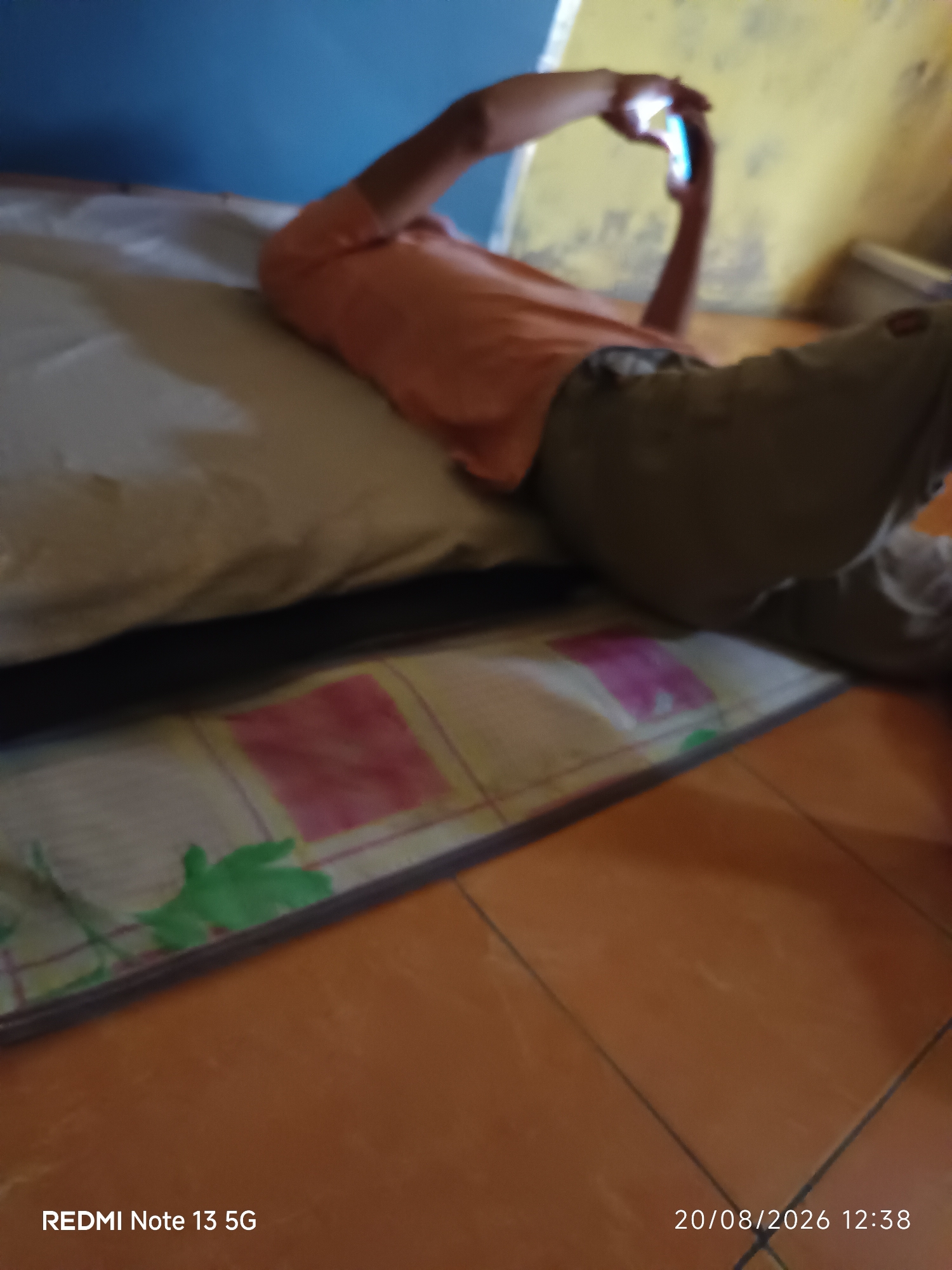 An individual is lying on their back on a mattress, propped up against a pillow, while holding a smartphone with their hands. The individual is wearing an orange shirt and grey pants. The scene appears to be indoors, with a blue wall in the background and a yellow wall to the right. The floor is tiled with orange tiles. The bottom right corner of the image displays the date "20/08/2026 12:38" and the text "REDMI Note 13 5G". There is no explicit information about the location being in Talaga Sari, Indonesia.