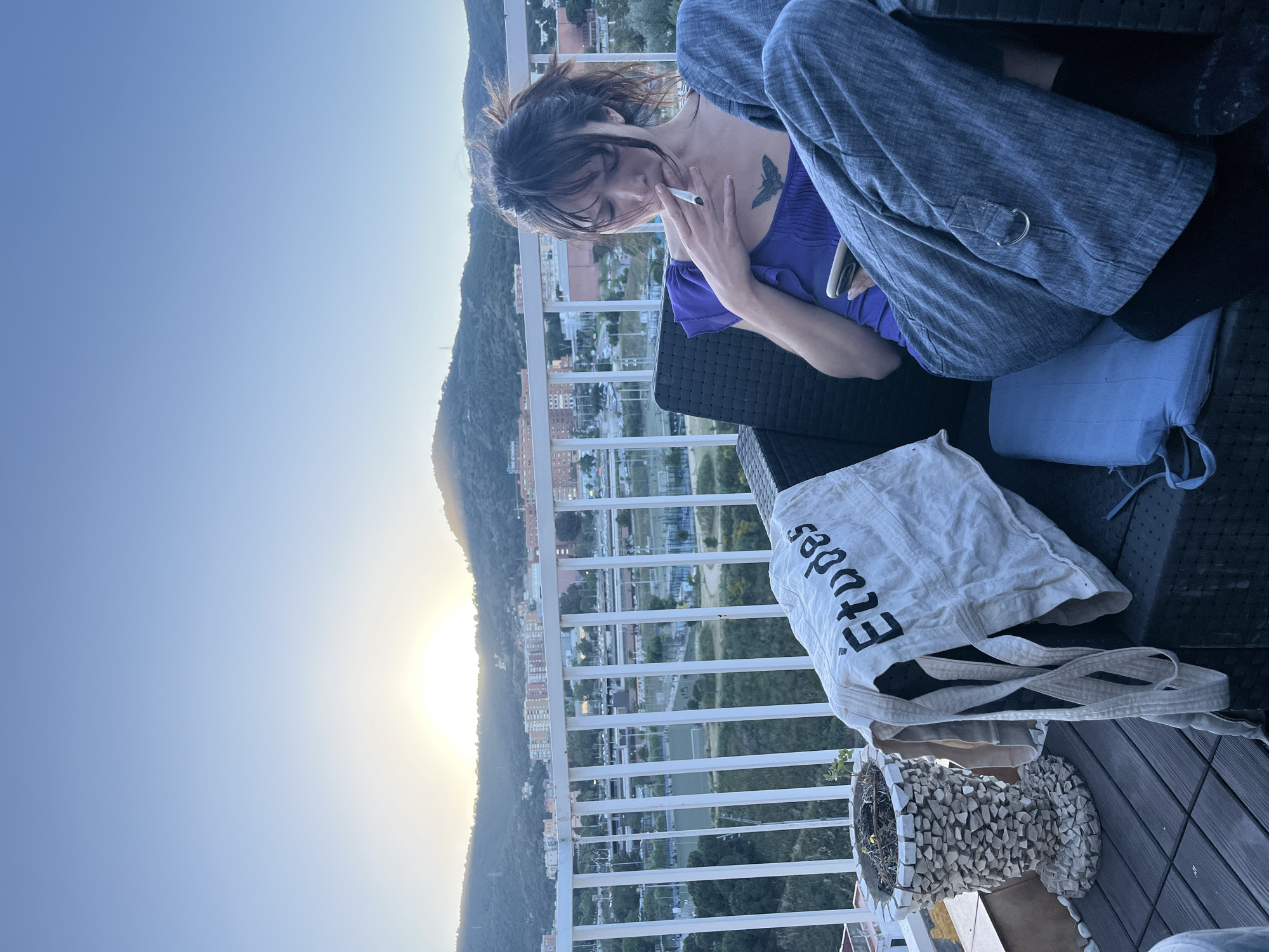 A woman is sitting on a balcony overlooking a cityscape in Barcelona, Spain, at sunset. She is smoking a cigarette and holding a phone in her other hand. She is wearing a purple shirt, a blue jacket, and black pants. A white tote bag with "études" printed on it sits next to her, along with a vase decorated with stones. The cityscape is visible in the background, with buildings and the setting sun illuminating the scene.