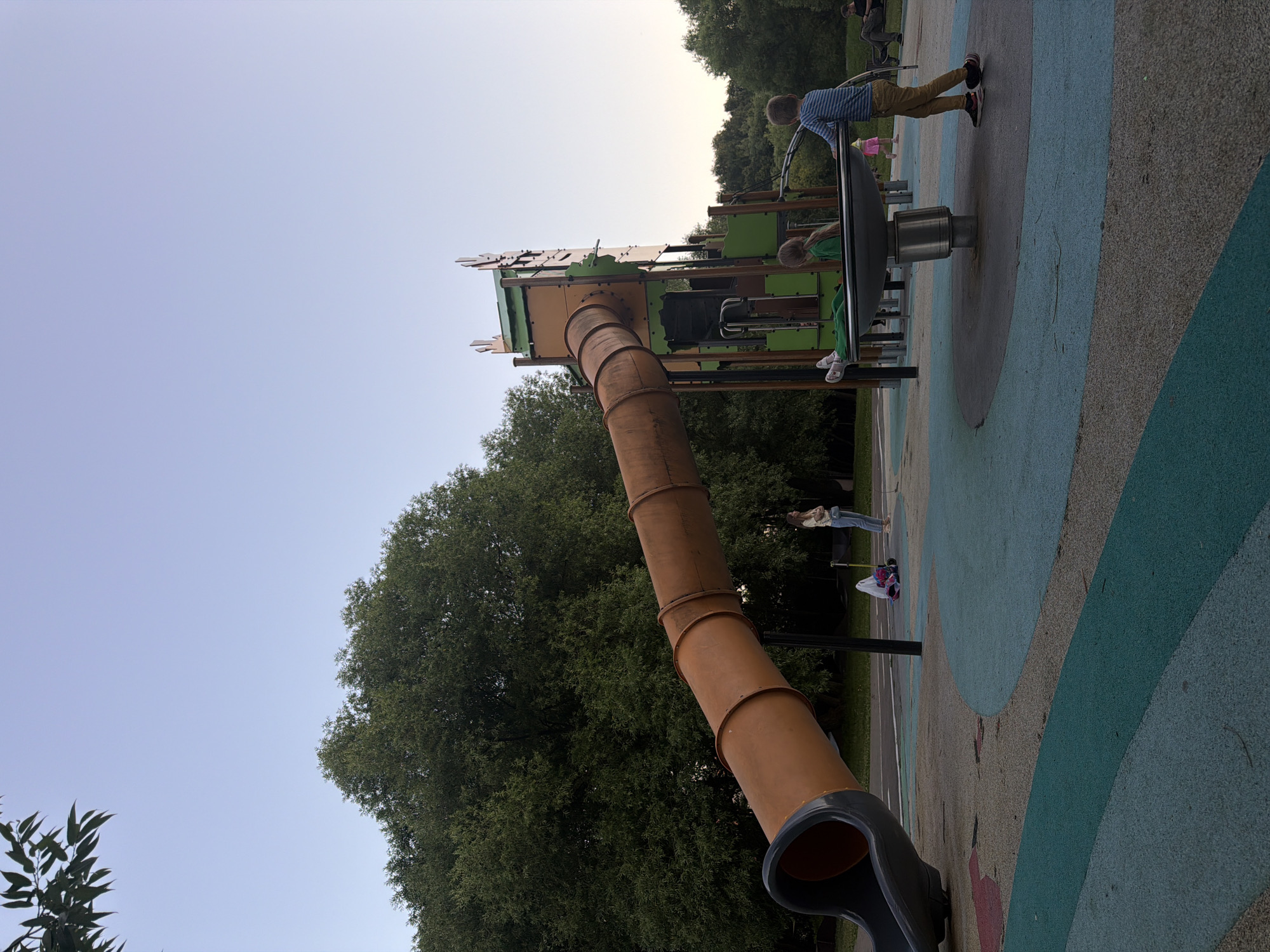The image depicts a playground scene on a clear day, with a large orange tube slide dominating the left side of the frame. In the background, a multi-level play structure with green and orange accents stands tall. Several children are present: one boy in a striped shirt and khaki pants is standing near a spinning apparatus, another child with long brown hair is seated on the same apparatus, and a smaller child in pink is visible further in the background. A woman in a light-colored top and jeans stands to the left of the play structure. The ground of the playground is a patterned surface of blue, grey, and beige. Large trees with dense foliage surround the playground. The sky is a pale blue, indicating it is either early morning or late afternoon. The location is identified as a playground in Moscow, Russia.