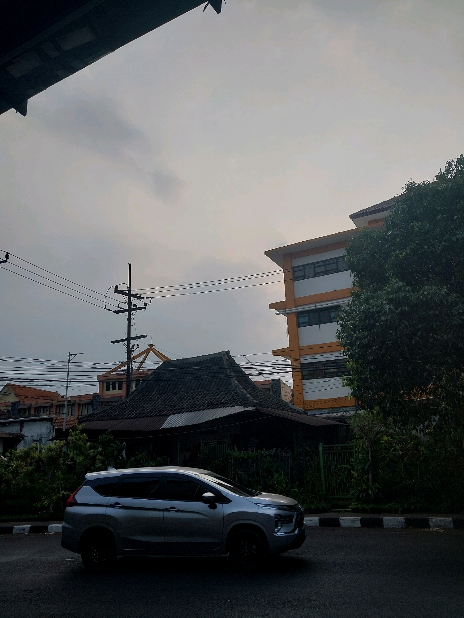 A silver minivan is captured in profile, driving on a paved road with a white and black striped curb. Behind the minivan, traditional Indonesian architecture with dark tiled roofs is visible, nestled amongst lush greenery. Further in the background, a multi-story building with orange and white facades stands, featuring numerous windows. Utility poles with a network of electrical wires crisscross the overcast sky. The scene is likely situated in Malang, Indonesia, given the architectural styles and the overall atmosphere of a Southeast Asian urban setting.