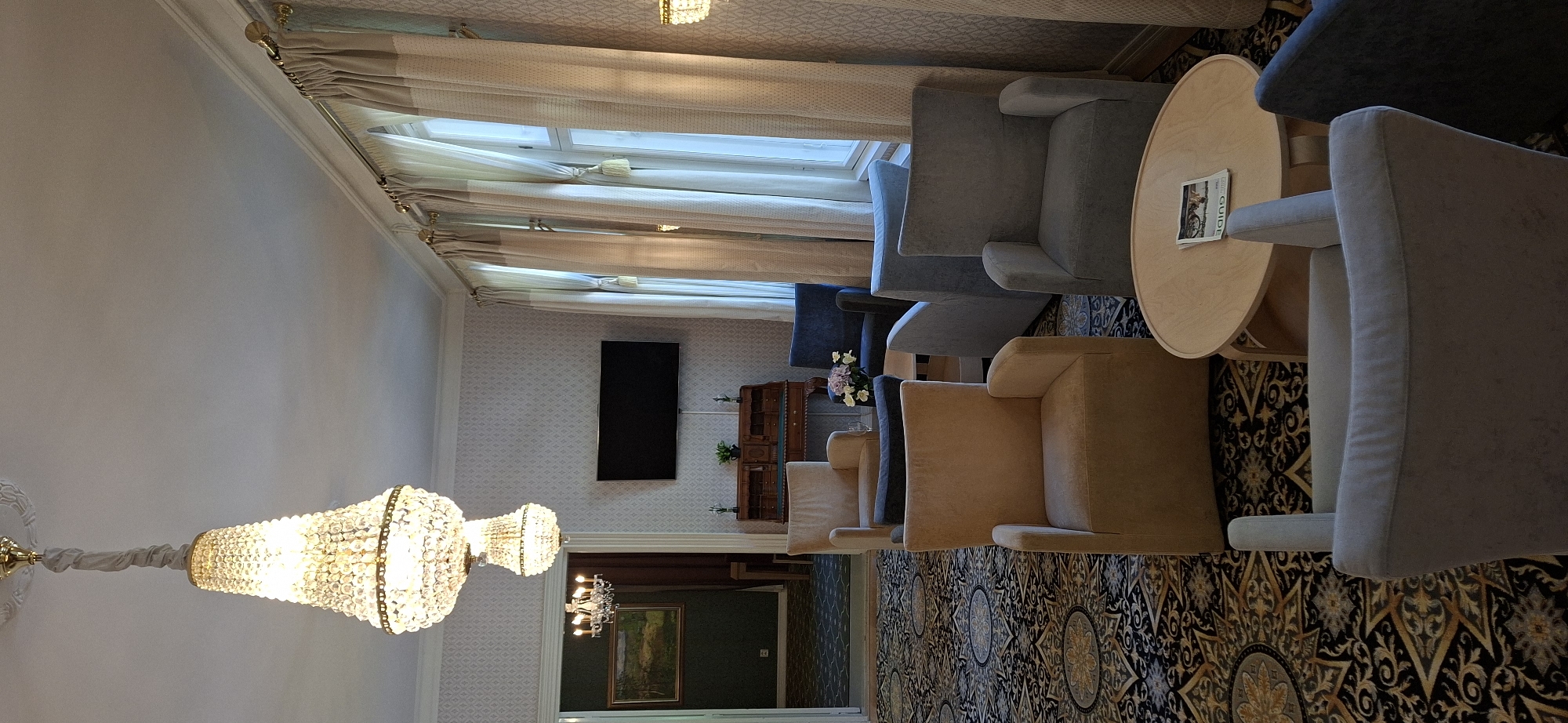 This image depicts an indoor scene, likely a lounge area, within the city of Lahti, Finland. The room is furnished with several upholstered armchairs in neutral tones, arranged around a light wood circular coffee table. The floor is covered with an ornate, patterned carpet. Two large crystal chandeliers hang from the ceiling. One chandelier is prominently visible in the foreground, while another, smaller one, is visible through an open doorway in the background, illuminating a hallway with a framed landscape painting on the wall. A flat-screen television is mounted on the patterned wallpapered wall. Behind the television, a wooden writing desk with drawers is visible. Large windows dressed with cream-colored curtains occupy a significant portion of the right wall. The overall impression is one of refined comfort, possibly in a hotel lobby or a similar public space. There are no people present in the image.