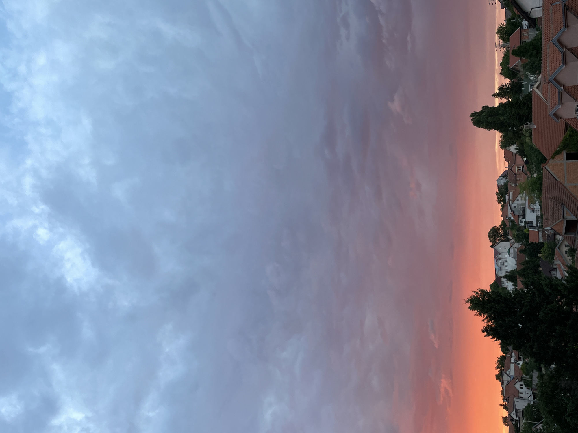 The image depicts a sunset over a residential area in Zvezdara Urban Municipality, Serbia. The sky transitions from a vibrant orange and pink hue at the horizon to a muted blue-gray with clouds towards the top of the frame. The foreground is dominated by the rooftops of houses, many with red-tiled roofs, nestled amongst lush green trees. The architecture suggests a mix of traditional and modern residential buildings. There are no people visible in the image. The overall scene conveys a sense of peaceful evening in a suburban setting.