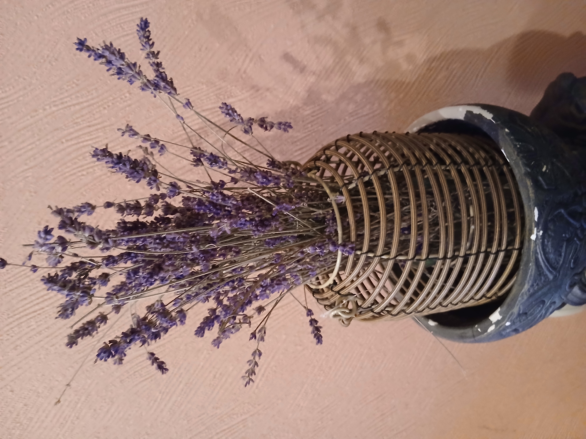 A bundle of dried lavender is displayed in a woven cylindrical vase with a metal frame. The vase sits inside a dark, ornate pot. The background is a textured, light-colored wall. The image was taken indoors.