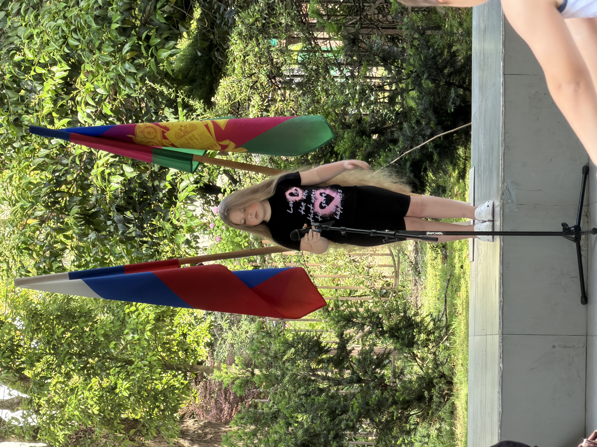 A young girl with long blonde hair is standing on a stage in front of two flags, one is the Russian flag, and the other is the flag of Krasnodar Krai. She is wearing a black t-shirt with text and graphics on it, and black shorts. She is holding a microphone in her right hand. The stage is outdoors, with trees and greenery in the background. There is a person in the foreground on the right, whose back is to the camera. The setting appears to be a park or an outdoor event space in Sochi, Russia.