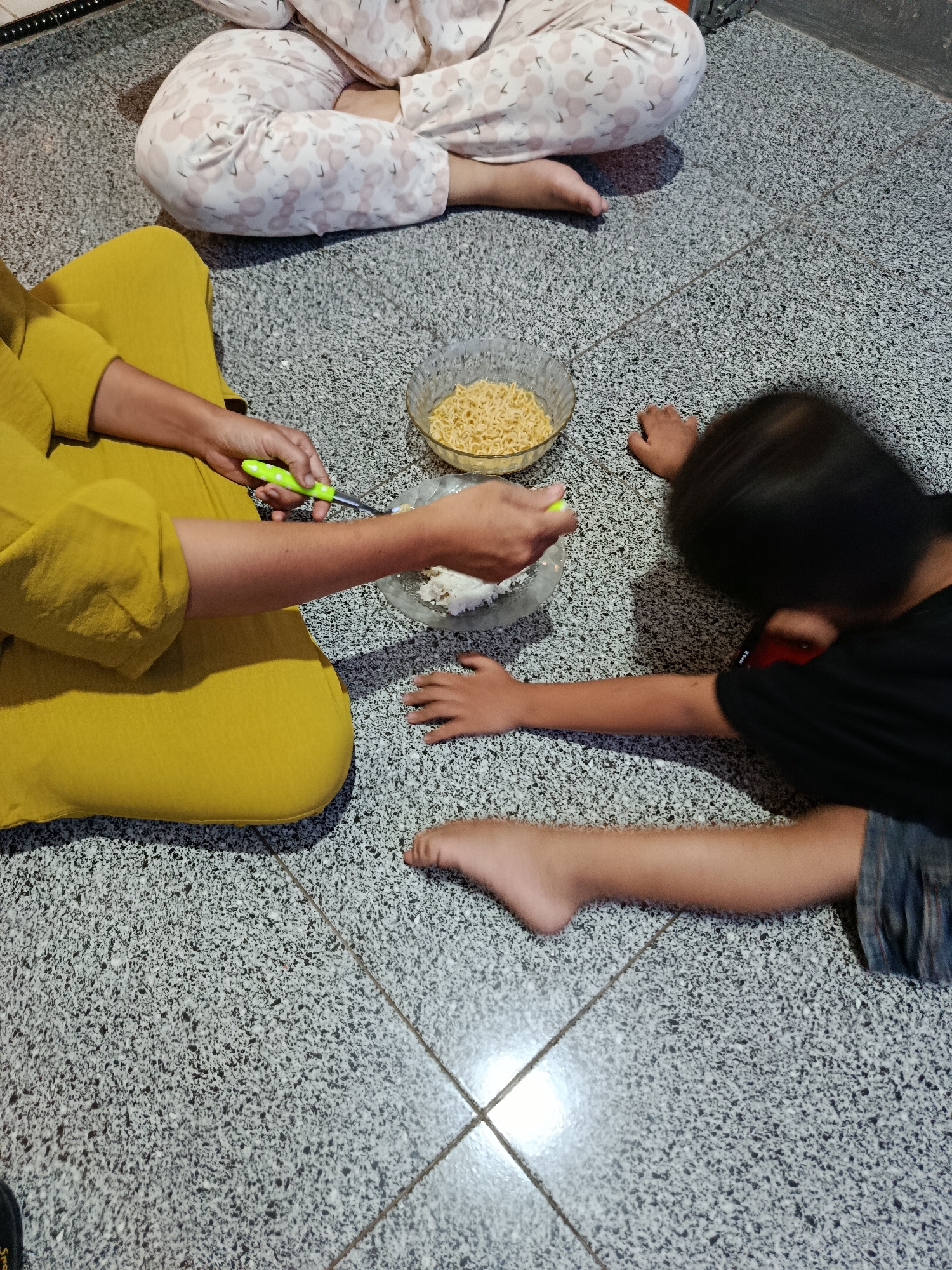 In Sarwadadi, Indonesia, a scene unfolds on a tiled floor where several individuals are seated or lying down. A person in a yellow garment is positioned in the foreground, holding a green-handled utensil over a plate of white food. To their right, a child in a black shirt and denim shorts lies on their stomach, reaching towards the plate. In the background, another person is seated, their legs covered in patterned pajamas. A bowl of instant noodles sits between the two seated individuals. The environment is a simple, indoor space with a speckled tiled floor.