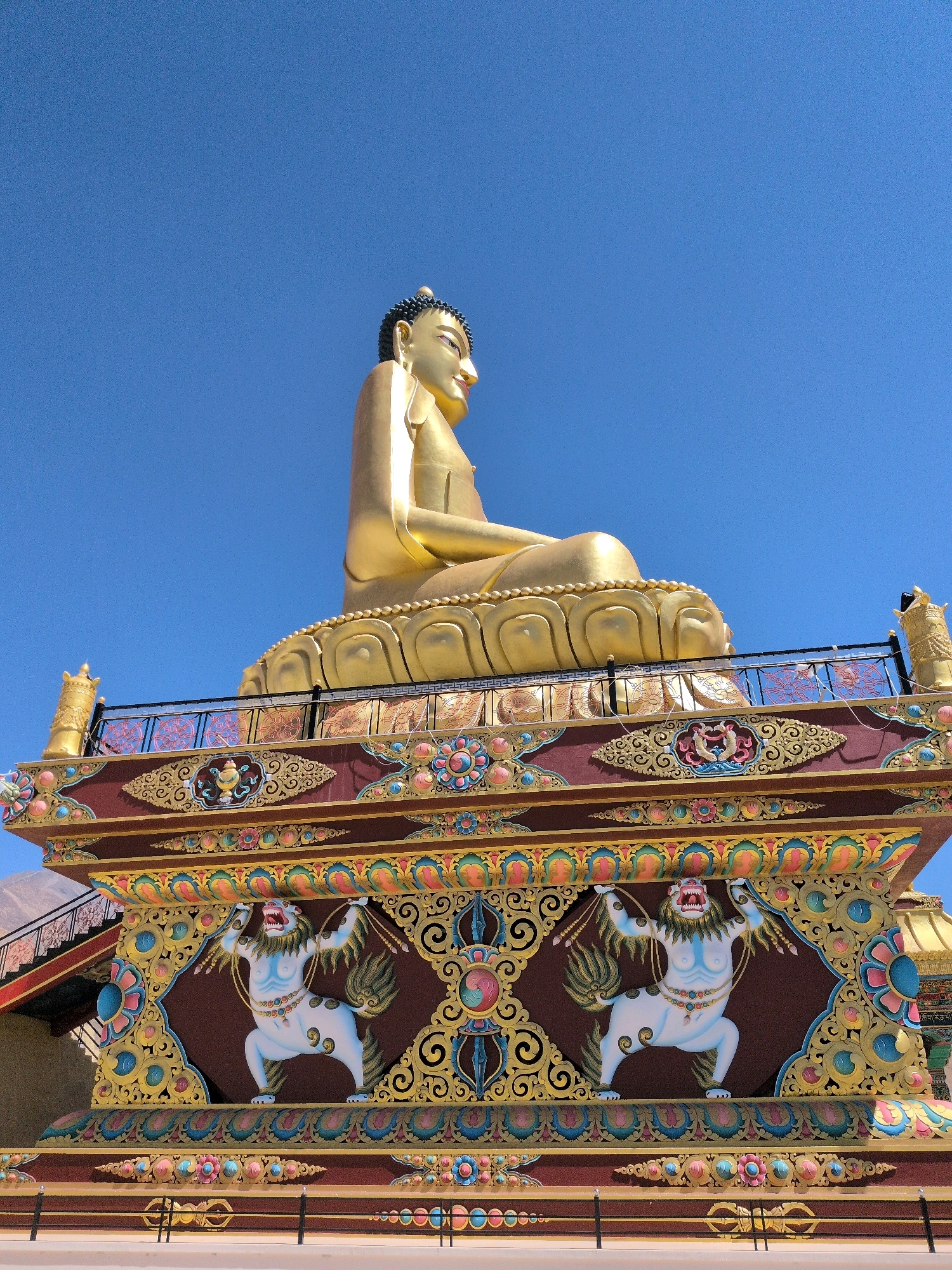 The image shows a large, golden Buddha statue seated on a multi-tiered pedestal. The pedestal is elaborately decorated with colorful patterns, Buddhist symbols, and carved figures of mythical creatures resembling lions. The statue is positioned against a clear blue sky. In the foreground, a staircase is visible leading up to the statue's platform. The overall scene suggests a religious or cultural landmark, likely a temple or monastery, in a bright, sunny environment. Based on the visual cues and context, this is likely the Maitreya Buddha statue located in Leh, India, a prominent Buddhist site.
