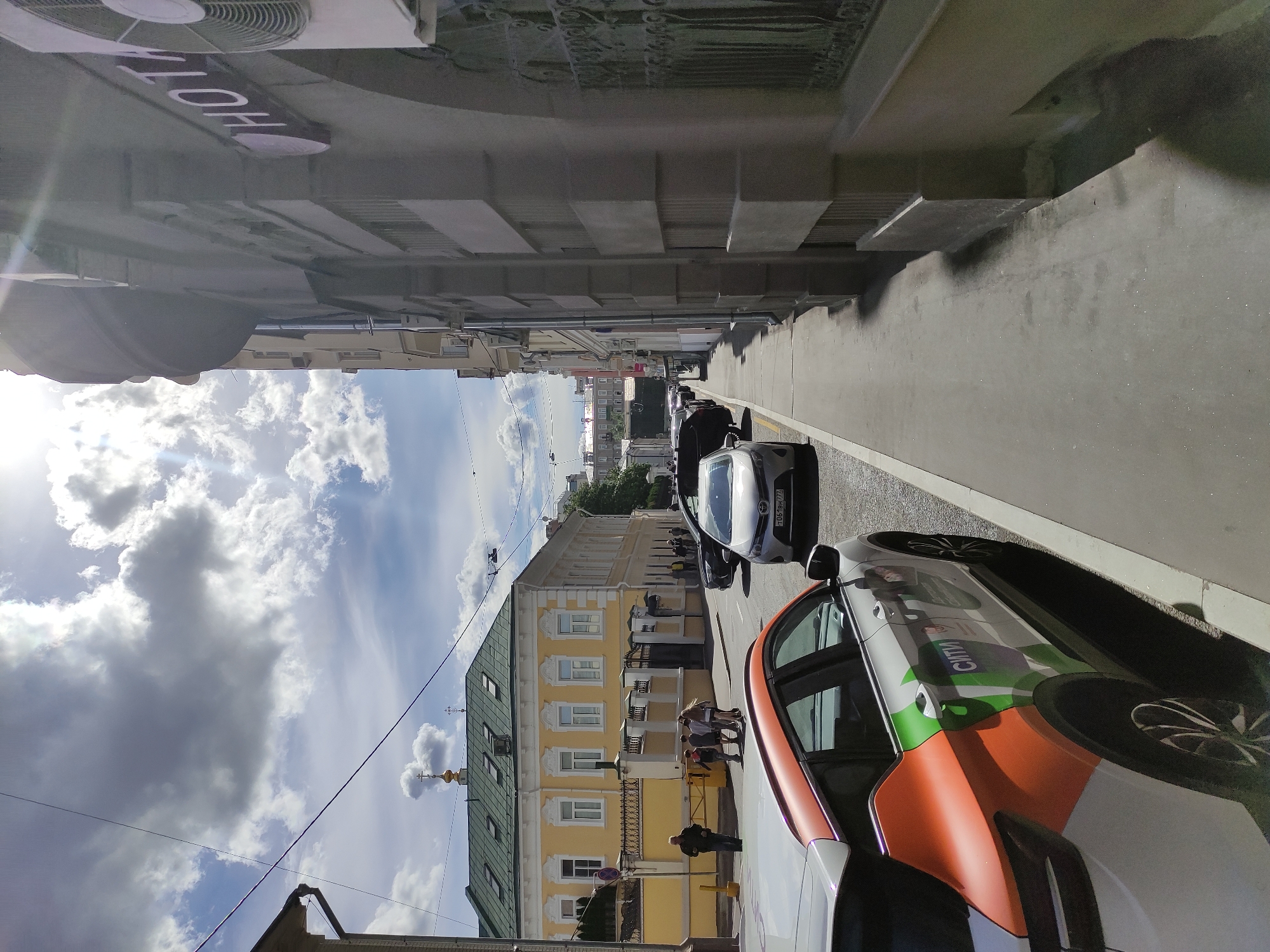 The image depicts a street scene in an urban environment. The location is likely Russia based on the Cyrillic characters on the vehicles. The view is of a street with parked cars on one side. The first car in the foreground is white with orange and green accents and the word "CITI" in Cyrillic. Adjacent to it is a gray car. In the background, there are people walking on the sidewalk and a building with a yellow facade and a green roof. The sky is visible with clouds and a few distant buildings. A sign with the letters "HOTEL" is visible in the top left corner. The perspective is from street level, and the image appears slightly rotated.