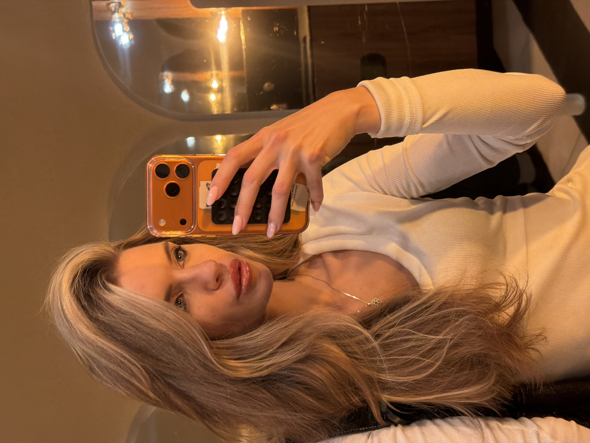 The image is a mirror selfie of a woman, oriented horizontally (rotated 90 degrees counter-clockwise from its original vertical capture). The setting appears to be indoors, under warm, artificial lighting, likely during the evening or night.
The primary subject is a woman with long, wavy, blonde and light brown highlighted hair. She is wearing a light-colored, possibly white or cream, ribbed long-sleeved top and a delicate silver necklace with a small, circular pendant. Her face shows a neutral to slightly alluring expression, with light eye makeup and prominent glossy lipstick.
She is holding an orange-cased smartphone in her right hand, positioned to capture her reflection in the mirror. The phone case clearly shows three camera lenses, characteristic of an iPhone Pro model. Her fingers, with manicured light pink or nude nails, are visible around the phone. A small, rectangular piece of white paper or sticker is tucked behind the phone within its clear case, displaying faint, indecipherable text.
The background, visible as a reflection in the mirror, is dimly lit with blurred, warm light sources, suggesting a cozy or intimate atmosphere. The surfaces reflecting these lights appear dark and glossy, possibly made of dark wood or metal. No specific location cues for Berlin, Germany, are visible within the image itself. There is no clearly readable text.