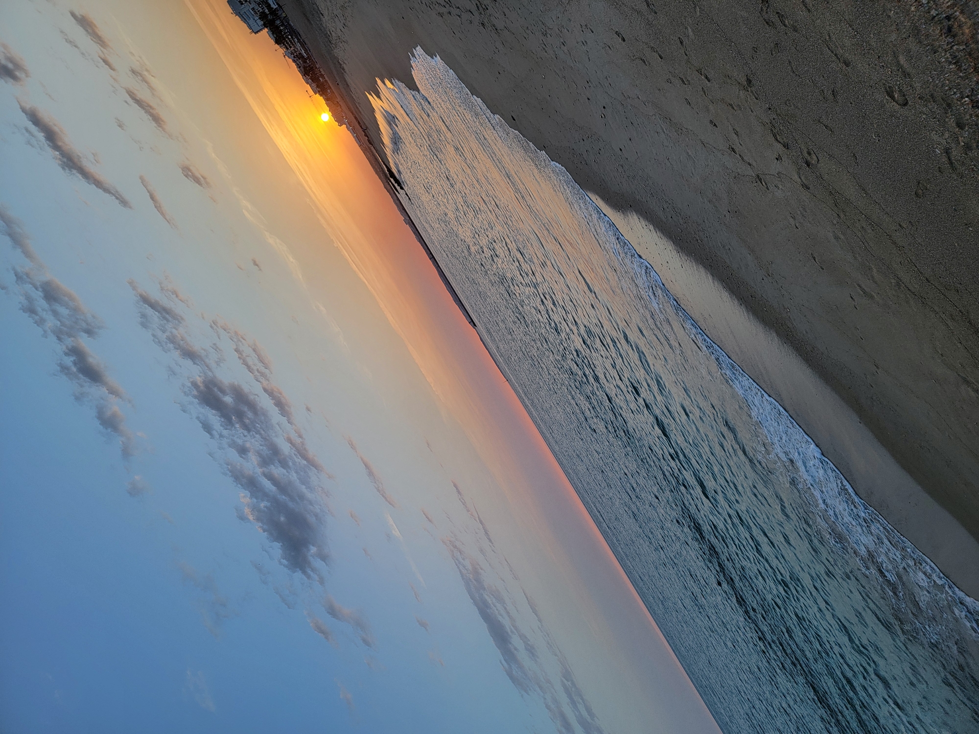 This is a photograph of a sunset at Quarteira Beach, Portugal. The image is taken from a low angle, looking diagonally across the sandy beach and the ocean towards the horizon.
The sky is a gradient of colors, transitioning from a clear blue at the top left to a soft pink and orange near the setting sun on the right. Wispy, elongated clouds are scattered across the sky, catching the warm light of the sunset.
The sun is a bright, golden orb low on the horizon, casting a radiant glow over the scene. In the background, along the coastline, are buildings that appear to be part of the Quarteira cityscape, silhouetted against the vibrant sky.
The ocean is calm, with gentle waves lapping at the shore. The water reflects the colors of the sunset, appearing in shades of blue, green, and orange. A thin line of white foam marks the edge of the receding tide.
The sandy beach occupies the lower right portion of the frame. The sand is a light brown color and shows numerous footprints, indicating recent human activity. The diagonal orientation of the shot adds a dynamic quality to the composition, emphasizing the expanse of the beach and the sea.
