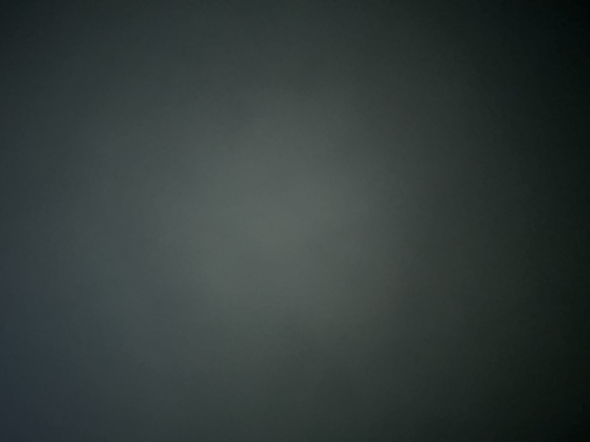 The image is a dark, abstract, and atmospheric shot, likely taken indoors or in low light conditions. It is predominantly a uniform dark gray with subtle variations in tone, suggesting a textured surface or a diffused light source. There are no discernible subjects like people, objects, or a clear scene. The activity or event, if any, is not evident due to the lack of clear visual information. Notable details such as time of day, weather, or location cues are absent. The overall impression is one of deep shadow and muted color. No text is visible in the image.