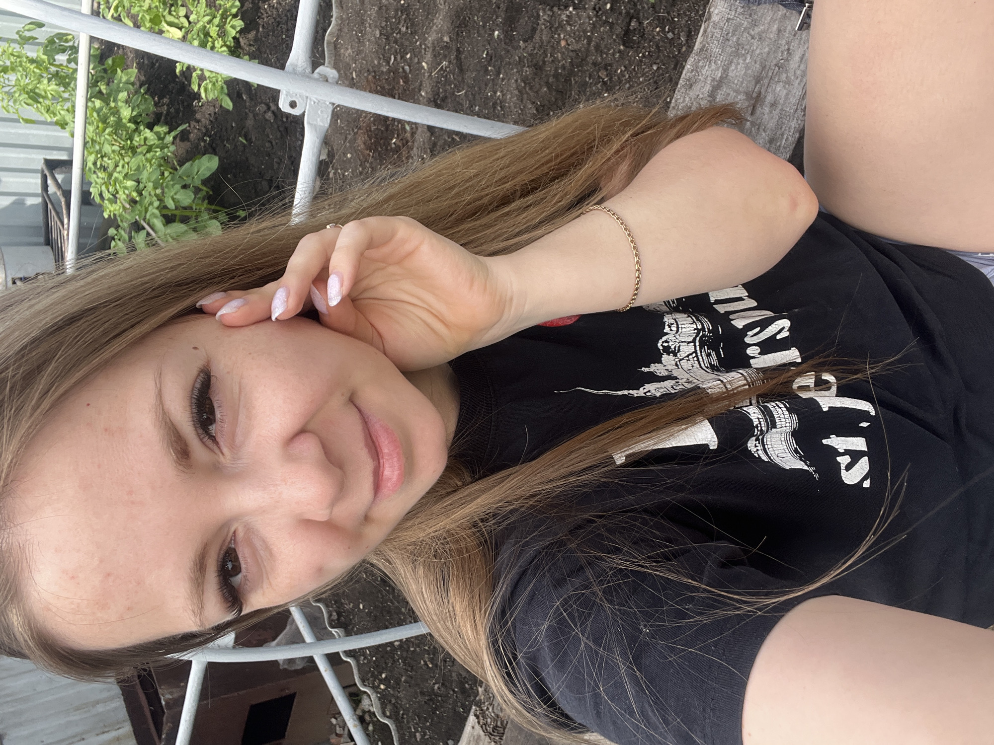 A young woman with long blonde hair is taking a selfie outdoors. She is wearing a black t-shirt with white text and an image of a city skyline. Her left hand is touching her face, and she has a gold bracelet on her right wrist. The background shows a garden with green plants and soil, surrounded by metal fencing. The image does not contain any specific visual cues to indicate the location is the city of Shadrinsk, Russia.