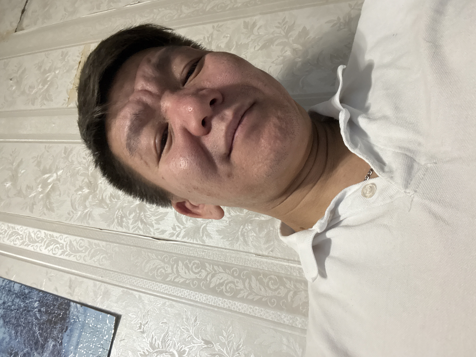 A low-angle, close-up shot features a man's face, appearing distorted due to the camera's angle. He has dark hair and is wearing a white polo shirt. The background consists of patterned wallpaper. A corner of a painting or framed image is visible in the lower left corner of the frame. The location is unknown.