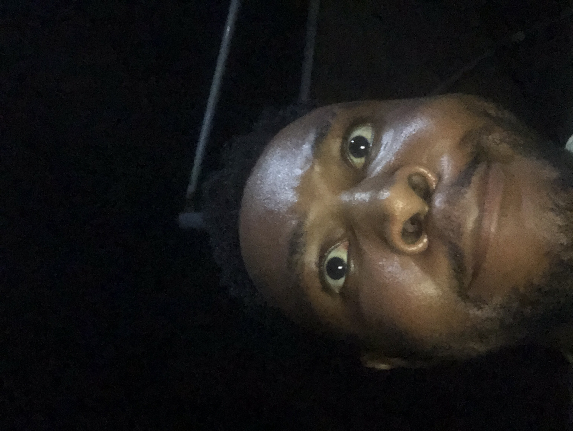 A close-up selfie of a dark-skinned man with visible moisture on his forehead and face. He is facing the camera at an odd angle, tilted to the right. The background is primarily dark, with the faint suggestion of vertical bars at the top of the frame. The setting is unknown.