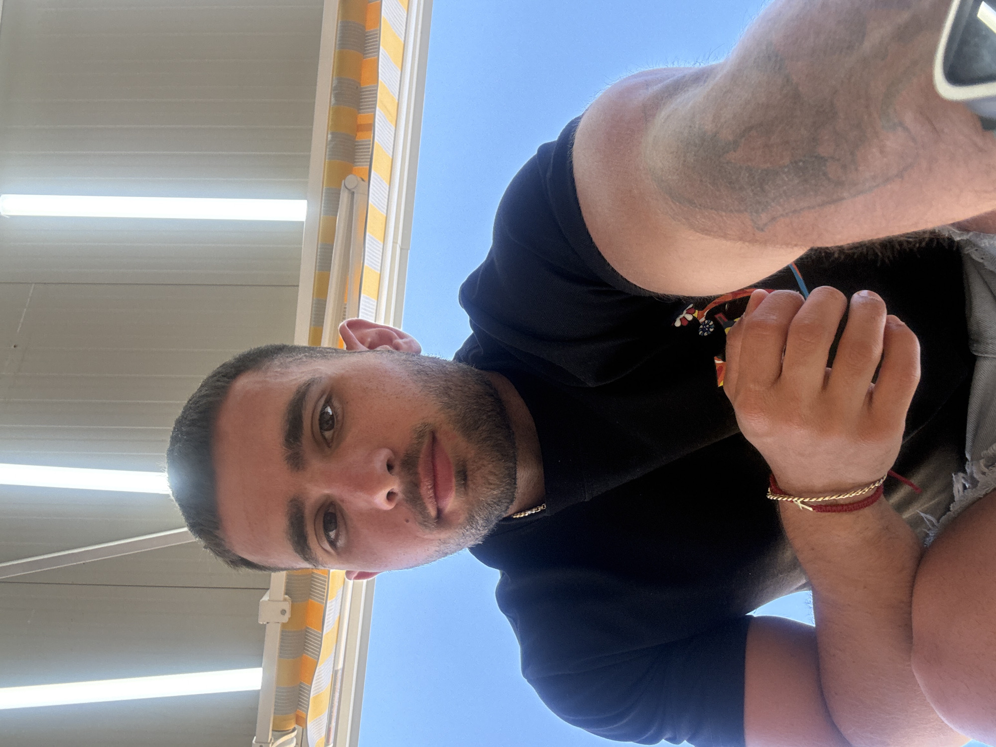 The image shows a close-up selfie of a young man with short dark hair and a beard, looking at the camera. He is wearing a black t-shirt and has a gold chain around his neck and two bracelets on his left wrist. He is outdoors, with a bright blue sky and a striped awning visible in the background. The awning appears to be made of alternating yellow and white stripes. The man is holding a small object in his right hand. The overall setting suggests a sunny day, possibly in a resort or outdoor dining area in Nesebar, Bulgaria, known for its coastal location and historical sites.
