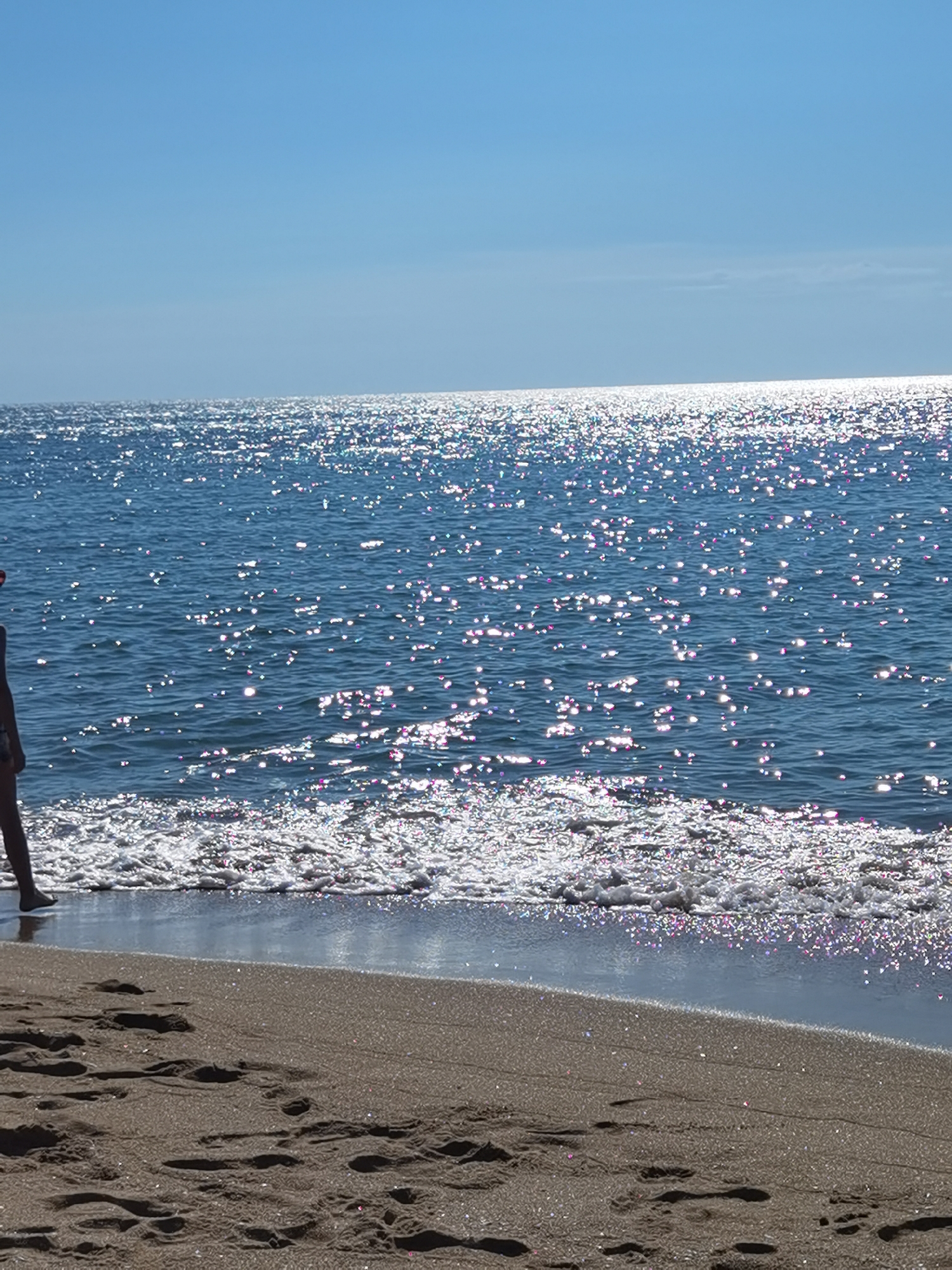 This image captures a serene beach scene under a clear blue sky. The sun glints brightly off the calm surface of the sea, creating a shimmering effect with highlights of pink and purple. Gentle waves lap at the shore, where the sand is imprinted with numerous footprints. In the foreground, a person is partially visible on the left, standing with their back to the camera, their legs and feet showing as they stand near the water's edge. The location is the beach in Varna, Bulgaria.