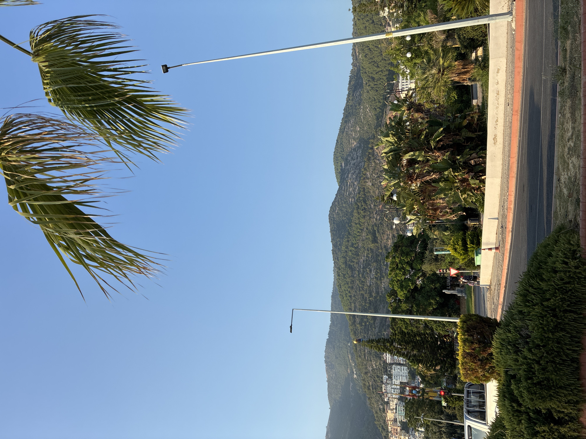 The image depicts a bright, sunny day in Alanya, Turkey, with a clear blue sky dominating the upper portion. In the foreground, the fronds of a palm tree sway gently, suggesting a breezy atmosphere. A tall, white lamppost extends upwards, its silhouette stark against the sky.
The midground features a steep, verdant mountain slope covered in trees. Below the mountain, a town with white buildings is nestled on the hillside. A road runs alongside the mountain, marked with a red and white striped curb. Another lamppost with a dark fixture stands near the road.
Further down, near the town, there are signs of activity. A traffic light is visible, with one red light illuminated. A white vehicle, possibly a van, is parked near a traffic light. A few people are gathered near a sign that reads "LUNA". Dense green bushes and trees line the foreground, providing a lush contrast to the urban and natural landscapes. The overall impression is one of a vibrant coastal town on a warm, sunny day.