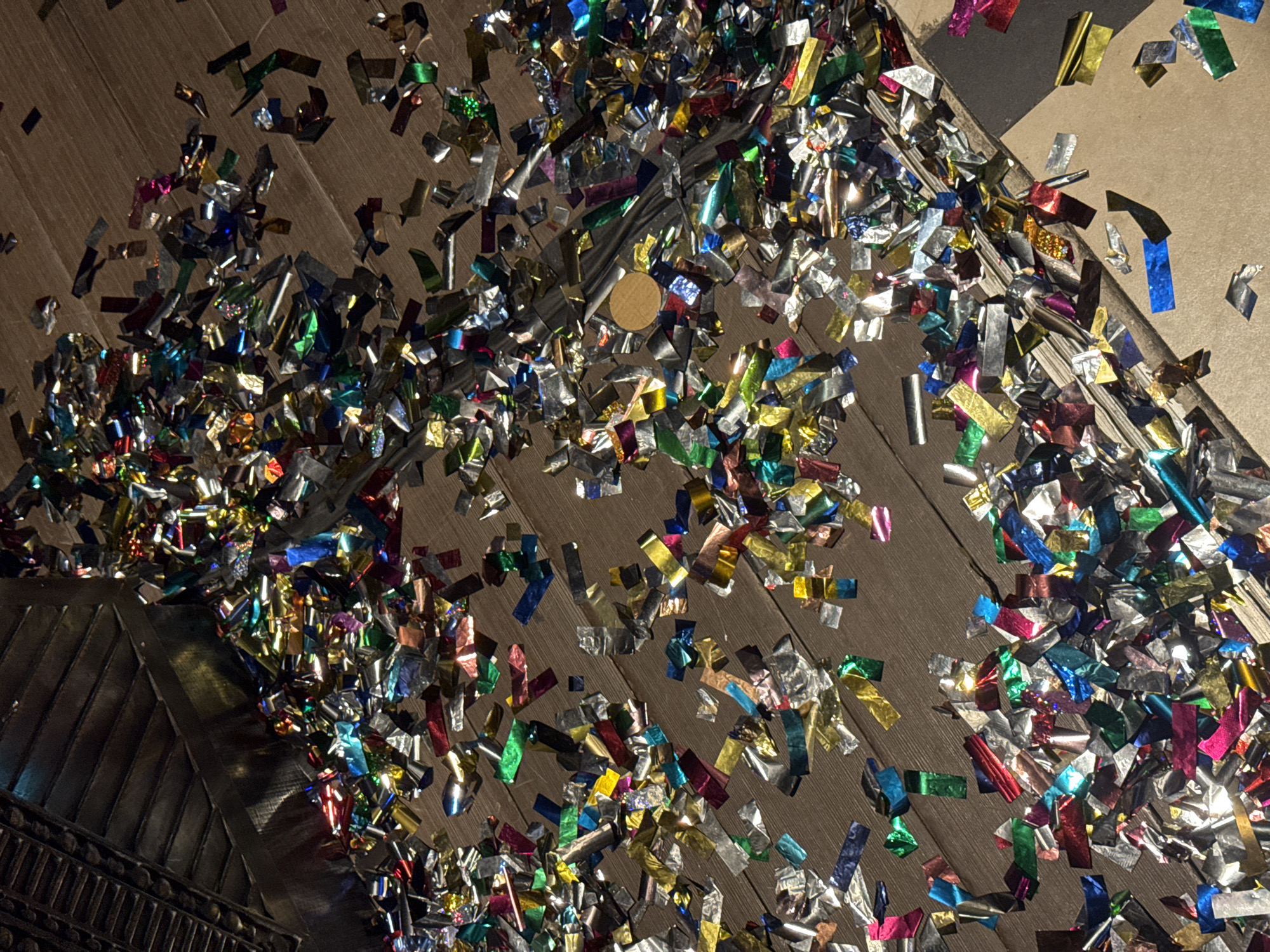 A high-angle shot depicts a wooden surface heavily covered in colorful metallic confetti of various shapes and sizes. The confetti is predominantly silver, gold, red, green, blue, and pink. There is a small round object (possibly a coin or token) visible amidst the confetti near the center of the image. The confetti appears to be concentrated in certain areas, leaving some sections of the wooden surface more exposed. A portion of an ornate metal structure is visible in the lower left corner. Based on the presence of confetti, the scene appears to be the aftermath of a celebration or event. The location (Dubai, United Arab Emirates) cannot be verified from the image alone.