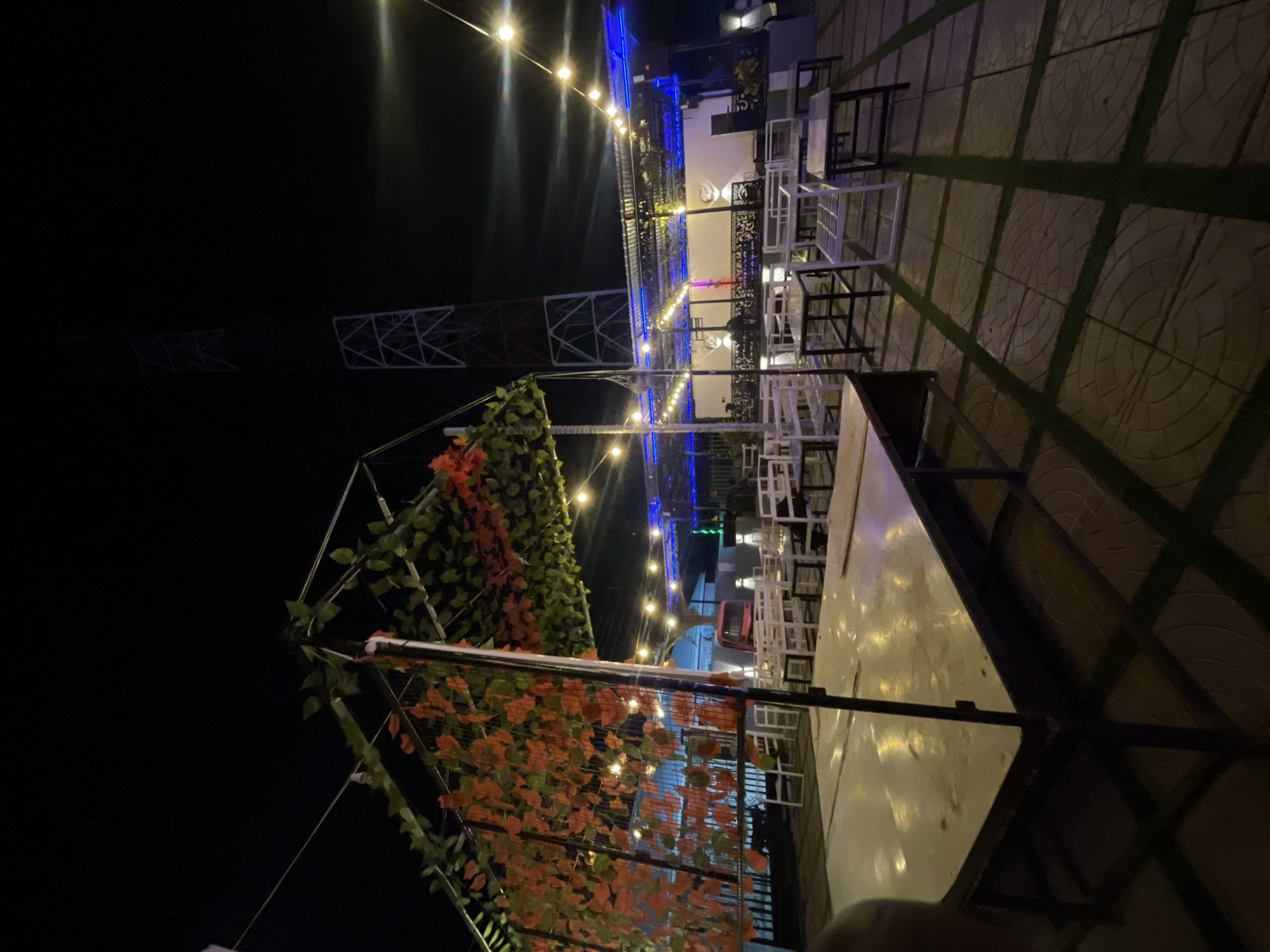 The image displays an outdoor dining or lounge area at night, captured from an elevated perspective and rotated ninety degrees clockwise. The scene, when viewed upright, reveals a sophisticated setup illuminated by numerous warm-toned string lights and cool blue accent lighting.
In the foreground, a trellis or pergola structure is visible, heavily draped with artificial green and reddish-orange foliage, creating a canopy effect. Below and to the right, a pristine white rectangular table is partially visible, suggesting a seating arrangement. The area features a mix of white and dark-framed tables and chairs with a modern, minimalist design, suggesting a contemporary bar or restaurant setting. The flooring consists of patterned light-colored tiles interspersed with strips of what appears to be artificial green turf or painted lines, forming a grid.
Further into the mid-ground, a multi-story building facade with distinct architectural features is lit with blue LED strips, adding a vibrant contrast to the warm string lights hanging above the seating areas. More string lights crisscross the space, contributing to the festive ambiance. In the distant background, against the dark night sky, the skeletal structure of a large tower or building frame is faintly visible, suggesting an urban or semi-urban environment. No people are present in the image.
The overall atmosphere is inviting and relaxed, characteristic of an evening spot designed for socializing or dining, consistent with the provided location context of "Bar Jirgi Summa, Nigeria." There is no visible text.