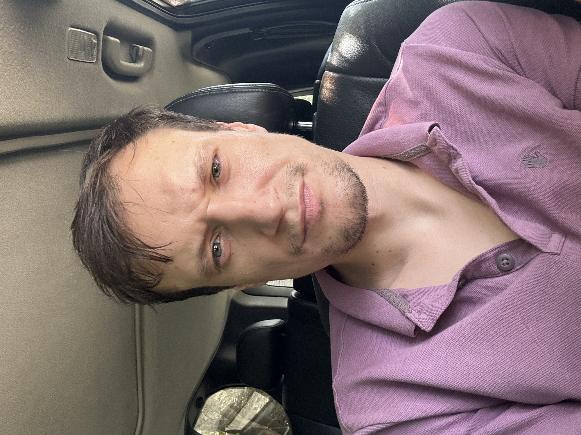 A man is seated inside a car, viewed from the chest up. He has short, dark hair and a light complexion. His expression is serious, with his brow furrowed. He is wearing a short-sleeved, purple polo shirt with a gray trim on the collar and a small, gray embroidered logo of a hand on the left chest. The interior of the car is visible, including the car's ceiling, a black headrest, and a black leather seat. Sunlight is filtering into the car, casting shadows. The man is looking directly at the camera. The image is a selfie taken from a slightly low angle. The location is identified as Makiv, Ukraine.
