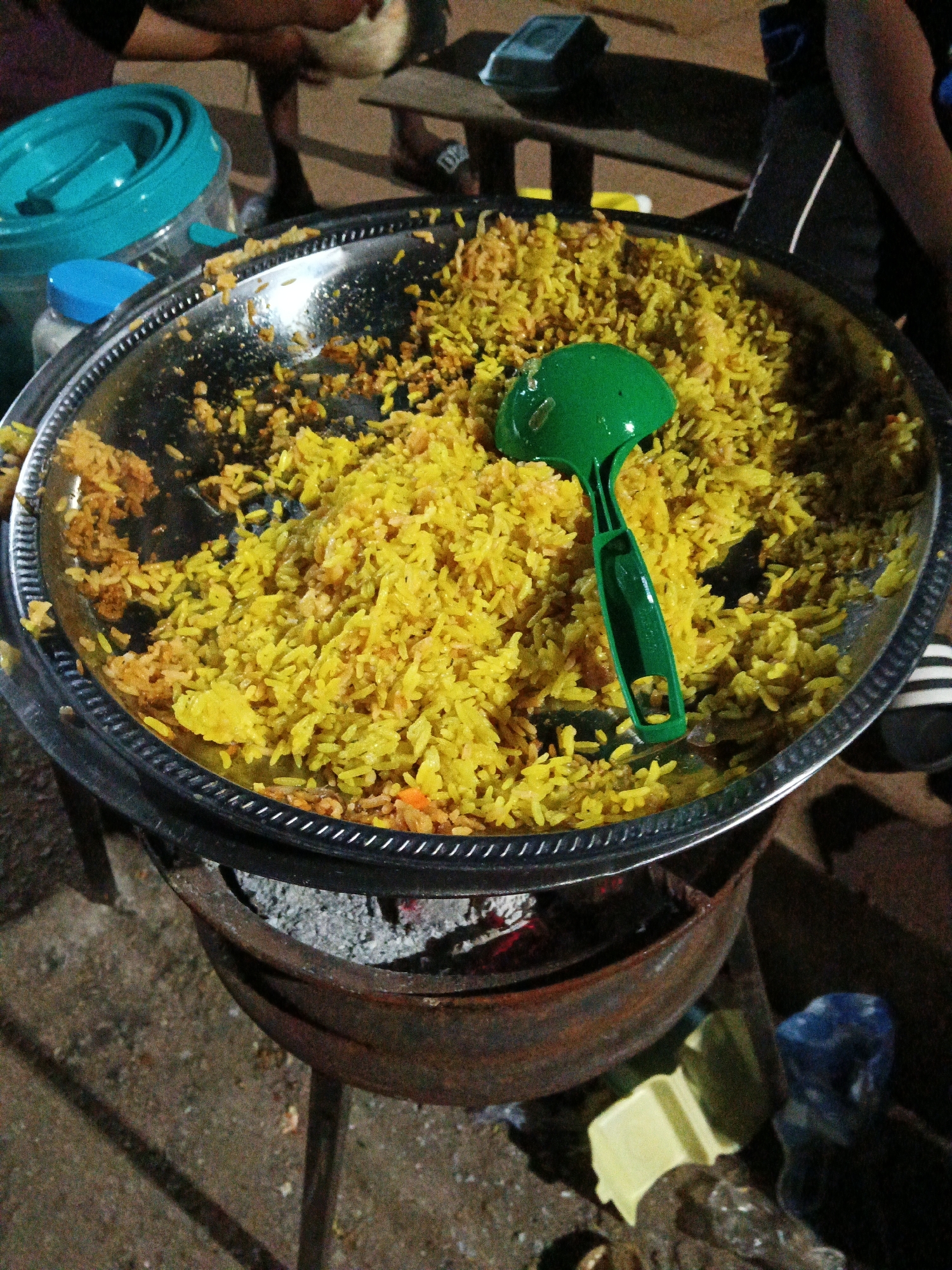 This image depicts an outdoor food stall in Bukuru, Nigeria, likely at night given the lighting and shadows. The main subject is a large, round metal tray filled with a generous portion of yellow-colored rice, possibly biryani or jollof rice, mixed with some visible pieces of orange. A bright green plastic ladle rests within the rice, suggesting recent serving or preparation. The tray is elevated on a rustic, dark metal stand, beneath which ashes and embers from a fire are visible, indicating the food is being kept warm or cooked over an open flame. To the left of the tray, a turquoise collapsible food container and a smaller blue-lidded container are present. In the background, out of focus, a person's legs and feet are visible, wearing sandals. The ground appears to be a mix of dirt and gravel. The overall scene suggests a busy, informal food vendor setting.