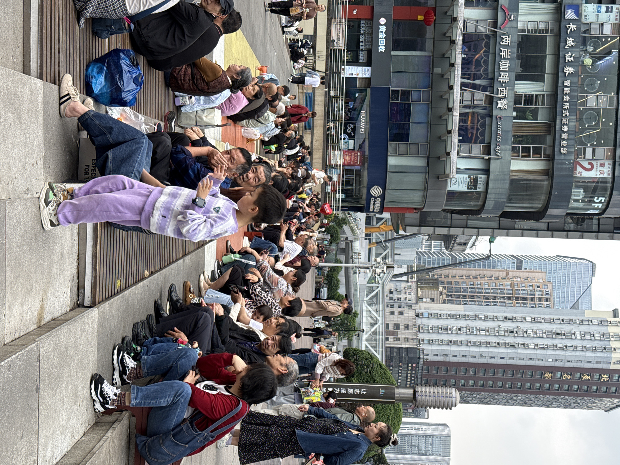 A crowd of people are sitting on a tiered structure of concrete and wood, gazing upward, possibly at a performance or event in the city of Liangjiang, China. The crowd is composed of a diverse mix of ages and attires, including individuals in casual wear, sportswear, and more formal outfits.
Many are resting on the steps, some using bags or cushions for comfort. A child in a purple tracksuit holds a phone, possibly recording the event.
Behind the seated crowd, a city street is visible, lined with tall buildings featuring various signs and advertisements in Chinese and English. These buildings include a Huawei store, a cafe named "Straits Cafe," and a financial institution, adding to the urban context of the scene.
The weather appears overcast, based on the sky conditions. A sign in Chinese is also prominently displayed, possibly indicating a point of interest or a direction within the city.