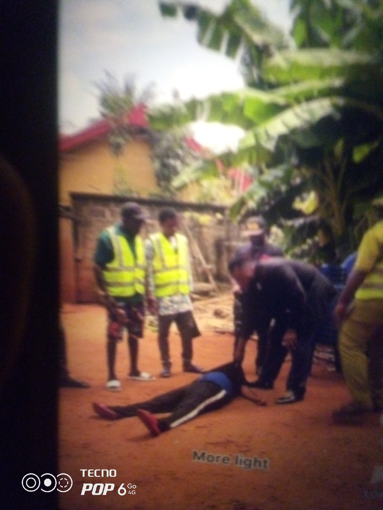 The image depicts an outdoor scene, likely a residential area, with a focus on a group of individuals gathered around a person lying on the ground. In the background, a yellow-colored building with a red roof is visible, along with lush green foliage, suggesting a tropical or subtropical environment. The ground appears to be dirt or sand.
There are at least five individuals in the frame. Two people in the middle ground are wearing bright yellow safety vests over dark shirts, with one wearing patterned shorts and the other patterned trousers. They appear to be observing the scene. To the right, another person in a yellow shirt is partially visible. In the foreground, a person is lying face down on the ground, wearing dark trousers with a white stripe and a blue undergarment. Their arms and legs are splayed. A few other individuals, dressed in dark clothing, are positioned around the person on the ground. One of them, wearing a dark jacket or top, is leaning over the person on the ground.
The overall impression is one of a situation requiring attention or assistance, possibly an accident or a medical emergency. The presence of people in safety vests might indicate they are involved in some form of community service or security. The image includes a watermark in the lower left corner with "TECNO POP 6 4G" and text that reads "More light" in the lower right corner. The resolution and focus of the image are low, leading to a blurred appearance of details.
