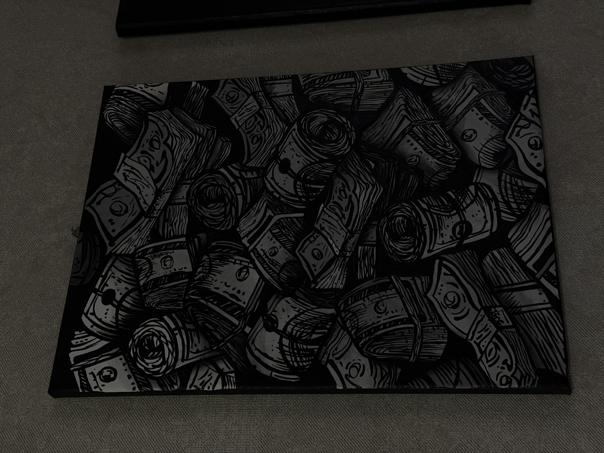 The media file presents an indoor view of a rectangular artwork displayed horizontally on a grey, textured surface, likely carpet or fabric. The artwork itself is a dark, monochromatic painting or print featuring a dense, stylized pattern of money bundles. These bundles are rendered with strong black outlines and varying shades of grey, depicting both stacks of banknotes and tightly rolled scrolls of currency. The style is graphic, utilizing hatching and stippling to suggest texture and form on the abstract money designs.
In the upper background, a dark, indistinct object, possibly another canvas or support, is partially visible. The overall lighting is dim and subdued, contributing to the dark aesthetic of the scene. There are no people or specific activities taking place. No discernible text or specific currency details are legible on the stylized money. The image context indicates the location is Chaikovsky, Russia.