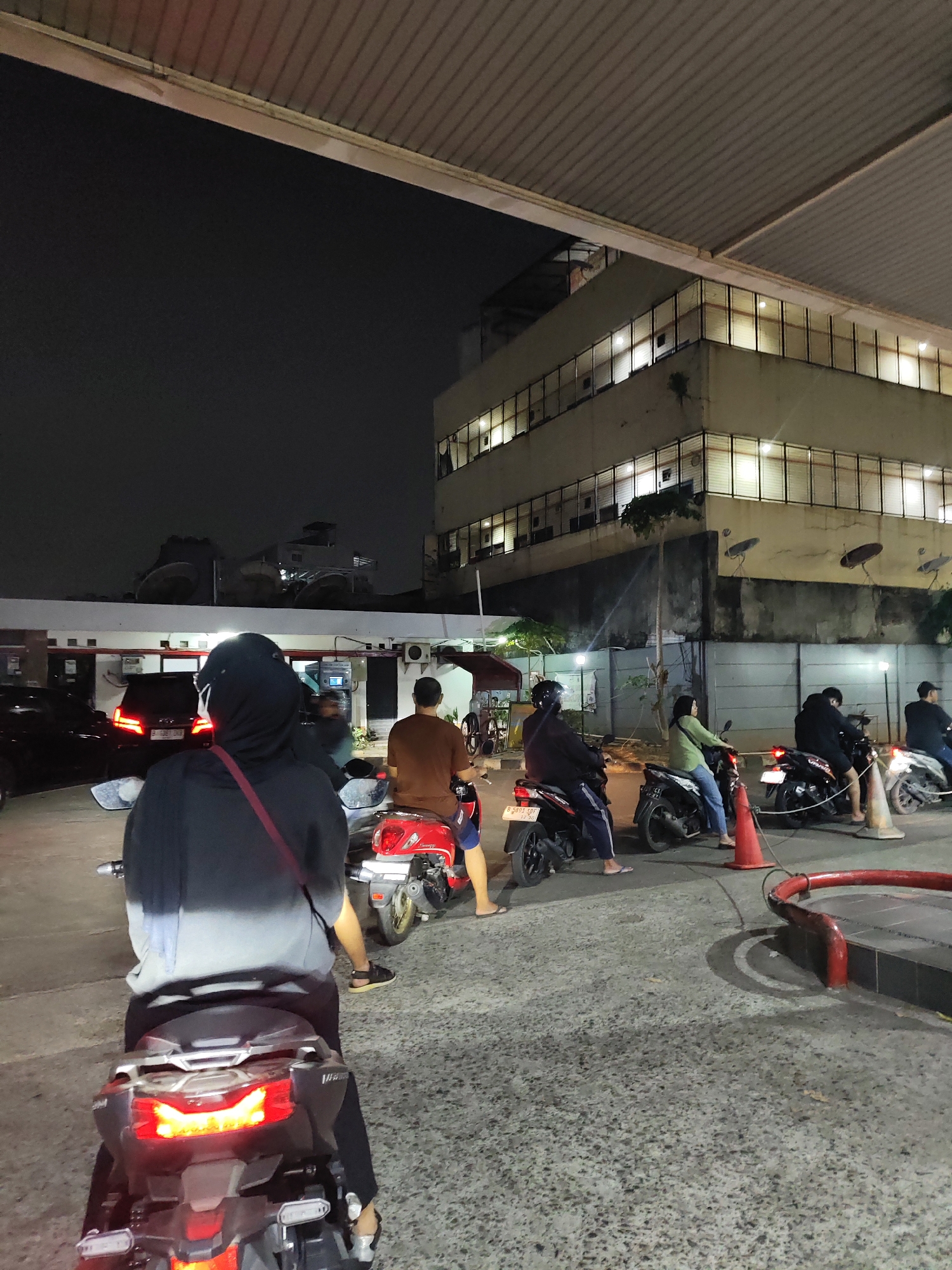 The image depicts a nighttime scene at what appears to be a gas station in South Jakarta, Indonesia. Several people are on motorcycles, waiting in line. In the foreground, a person is seated on a dark-colored scooter, facing away from the camera. Behind them, a red scooter is visible with another person seated on it. Further back, more motorcycles and riders are lined up, some with helmets on. A multi-story building with illuminated windows stands in the background, and a portion of a covered roof structure is visible at the top of the frame. The ground is asphalt, and orange traffic cones are present. The lighting suggests it is evening or night, with artificial lights illuminating the area.
