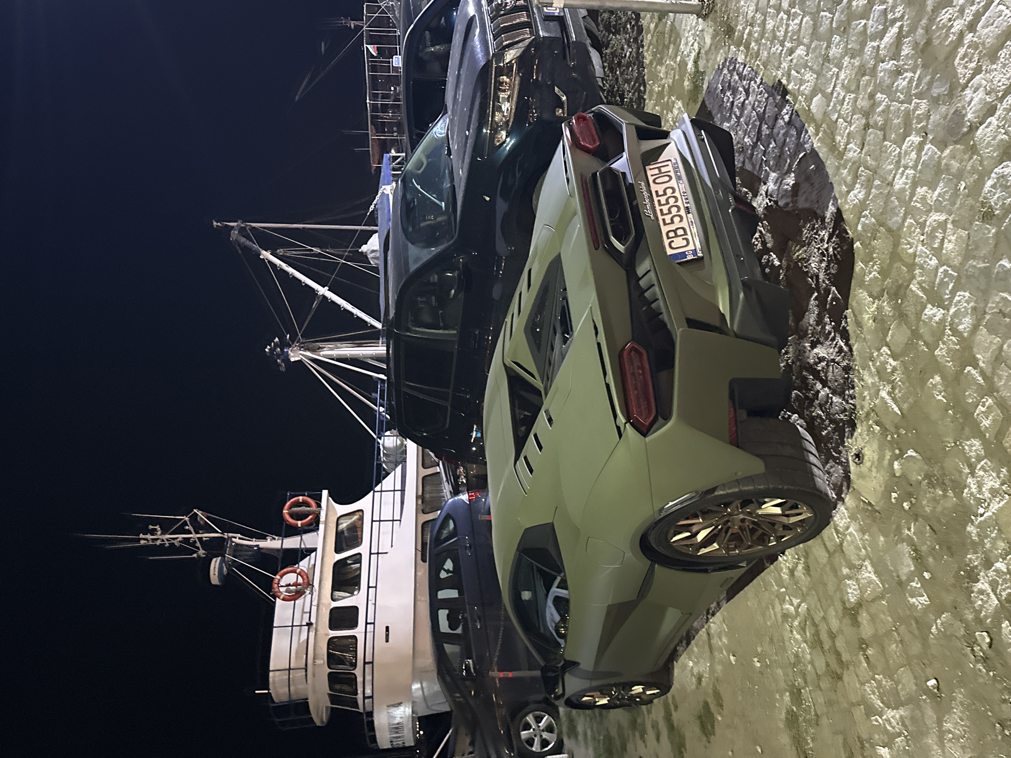 In the city of Sozopol, Bulgaria, at night, several vehicles are parked on a cobblestone surface near a large white boat docked at a harbor. The foreground is dominated by a matte green Lamborghini, its rear angled towards the viewer. The car has gold-colored wheels and a license plate that reads "CB 5555 OH". Behind the Lamborghini, a dark-colored SUV, possibly a Toyota Land Cruiser, is parked. Further back, a blue hatchback and a darker car are also visible. The white boat, which appears to be a passenger vessel, is illuminated and features two orange life preservers on its railing. The sky is dark, indicating nighttime. The scene is likely a waterfront area in Sozopol, possibly a quay or parking lot adjacent to the harbor.