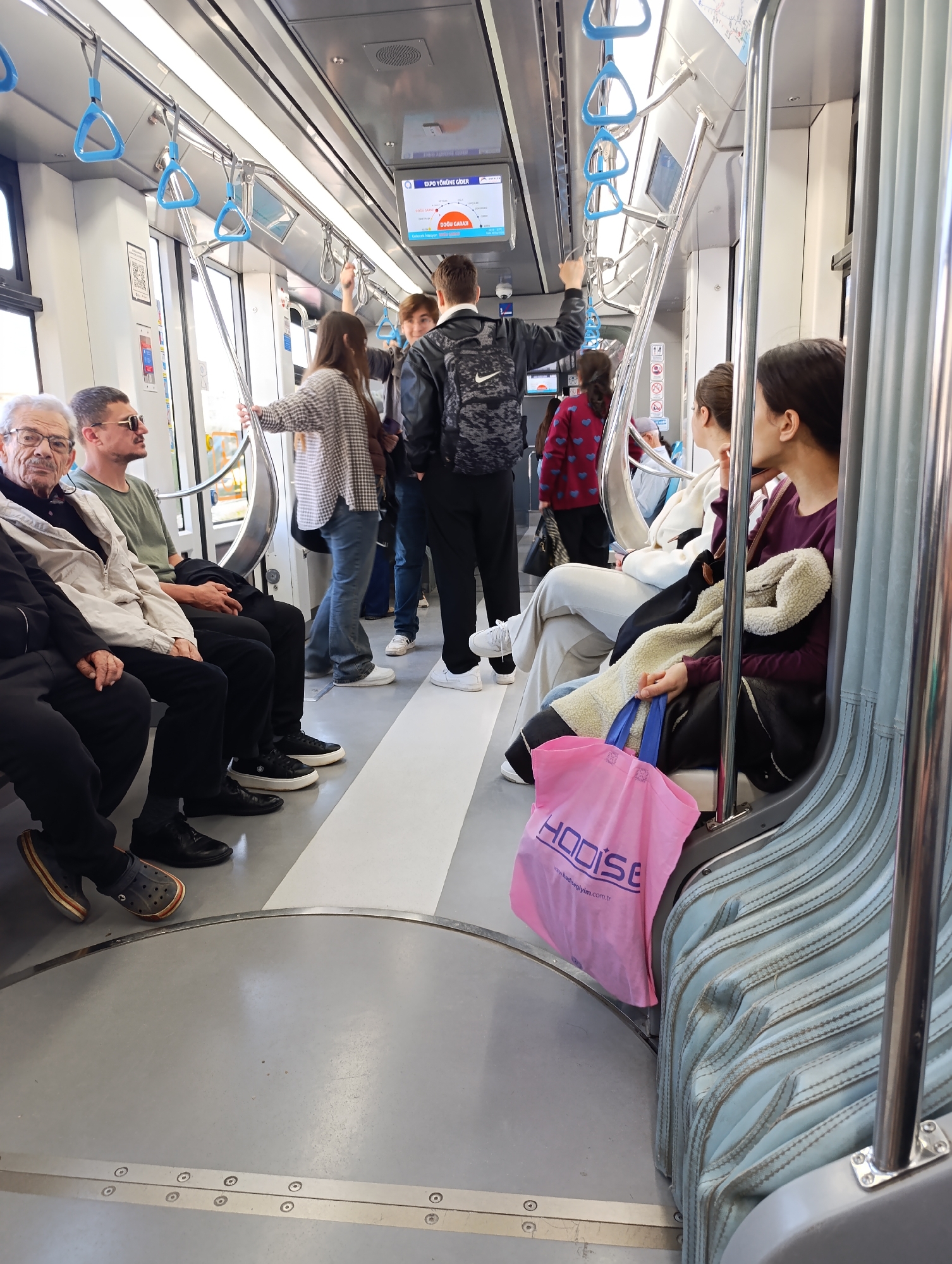 The image shows the interior of an Antalya, Turkey subway car, with multiple people present. Several passengers are seated on blue, ribbed seats, with one person holding a pink shopping bag with the word "HODISE" printed on it. Other passengers are standing, some holding onto the blue hanging straps. The floor is gray with a white stripe down the middle. Some passengers are wearing casual clothing like jeans, sweaters, and sneakers, while others wear jackets. There's a screen visible near the ceiling displaying a train route map.