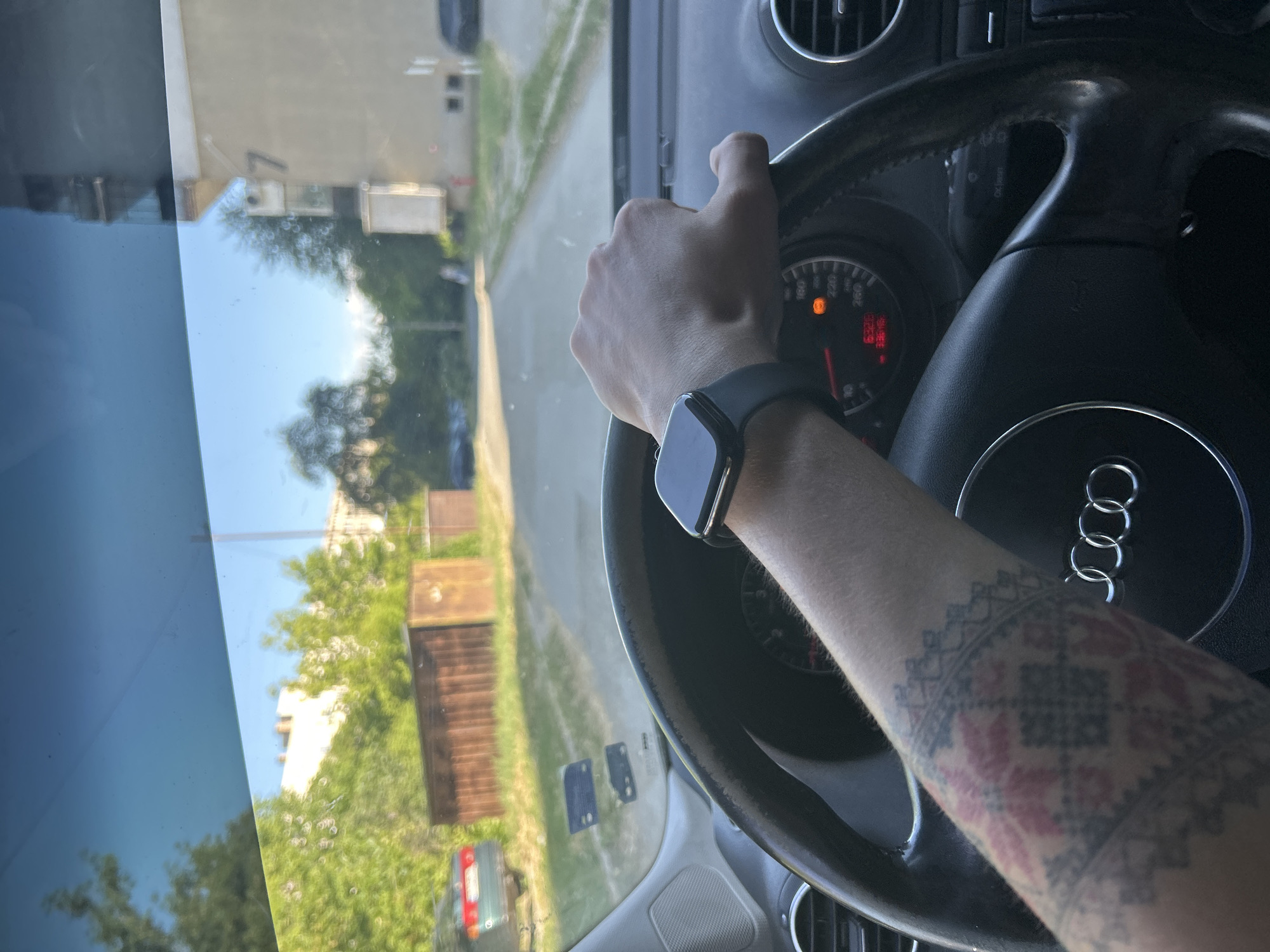 A person is driving a car, presumably an Audi, as indicated by the steering wheel logo, through a residential area. The driver is wearing a smartwatch on their left wrist. The location appears to be Kazanlak, Bulgaria, judging from the buildings visible through the windshield, and the style of the tattoo visible on the driver's arm. The car's dashboard is visible, showing the instrument cluster and air vents. The outside environment includes buildings, trees, and a road. The car is presumably moving as indicated by the blurred background.