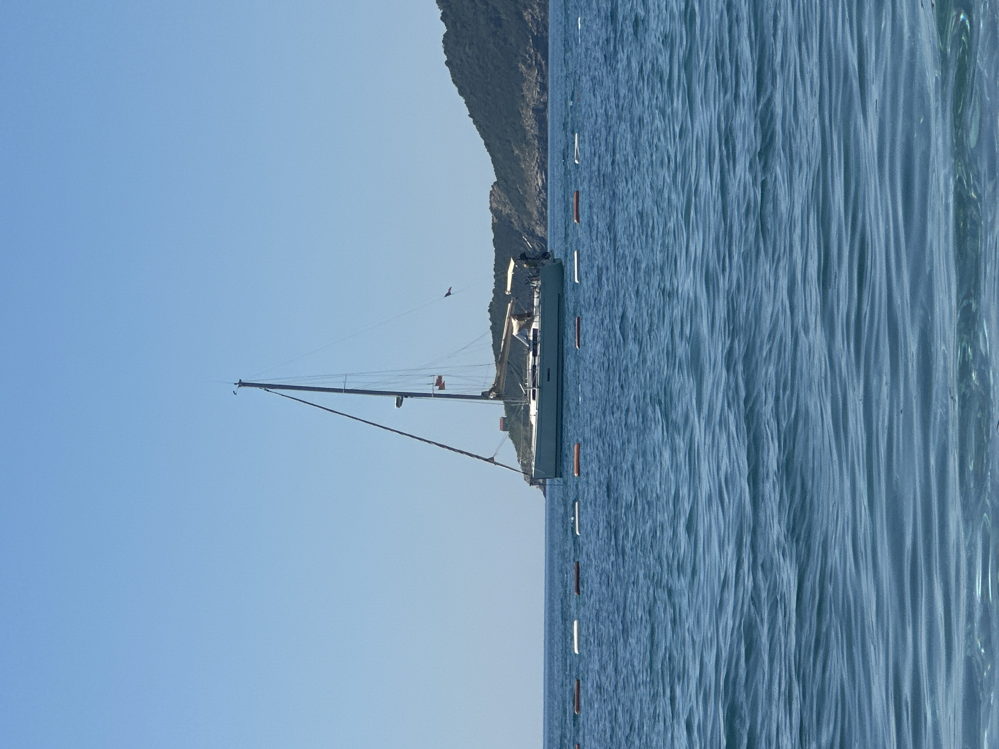 A sailboat is anchored offshore in Boreti, Montenegro. The view includes a clear blue sky, calm blue water, and a mountainous coastline in the background. Buoys line the water.