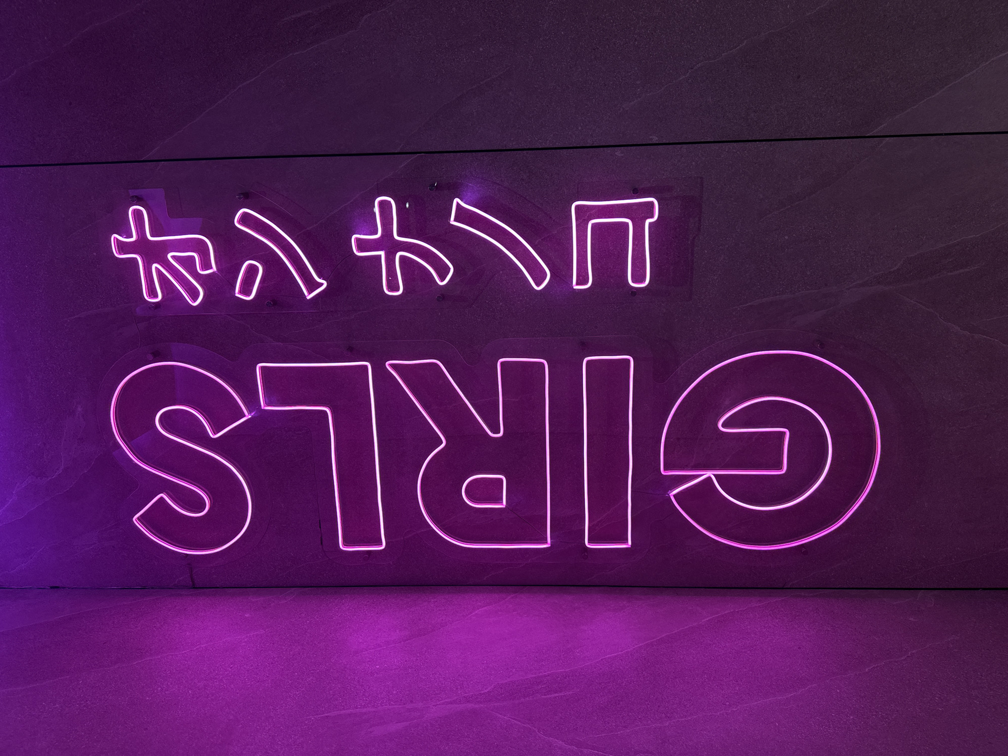 A purple neon sign spelling out "GIRLS" in English is mounted on a dark, mottled wall. Above it are four Japanese katakana characters, also rendered in purple neon. The scene is illuminated by the sign itself, which casts a purple glow onto the floor. The wall and floor surfaces appear to be made of a stone-like material. Based on online search results, this sign can be found in North Kuta, Indonesia, within the vicinity of the bar 40 Thieves.