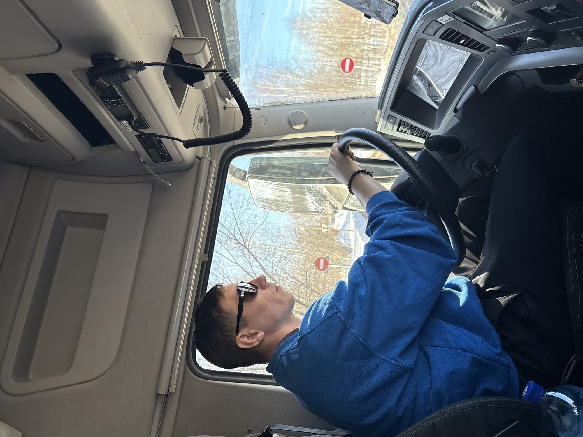 A male individual is seated in the driver's seat of a truck cab in Staroe Mikhailovskoe, Russia, operating the vehicle. He wears a blue long-sleeved top, black trousers, reflective sunglasses, and a dark beaded bracelet on his left wrist. His left hand is positioned on the black, textured steering wheel, and his head is turned slightly right, looking forward through the windshield.
The cab interior features a light gray overhead console containing what appears to be communication equipment with two coiled microphone cords and handsets, along with integrated storage compartments. The dashboard to the driver's right displays an illuminated digital screen, air vents, and various control buttons. A plastic water bottle with a blue cap is located in the lower left of the frame, near the seat. A mobile device mount is partially visible on the far right of the dashboard.
The exterior environment, viewed through the windshield and side window, depicts a winter landscape. The ground is covered with snow, and deciduous trees without leaves line the background under a clear blue sky. Two distinct red and white circular "no entry" road signs are visible on posts along the roadside. The truck's side mirror is also visible on the left, reflecting the snowy road.