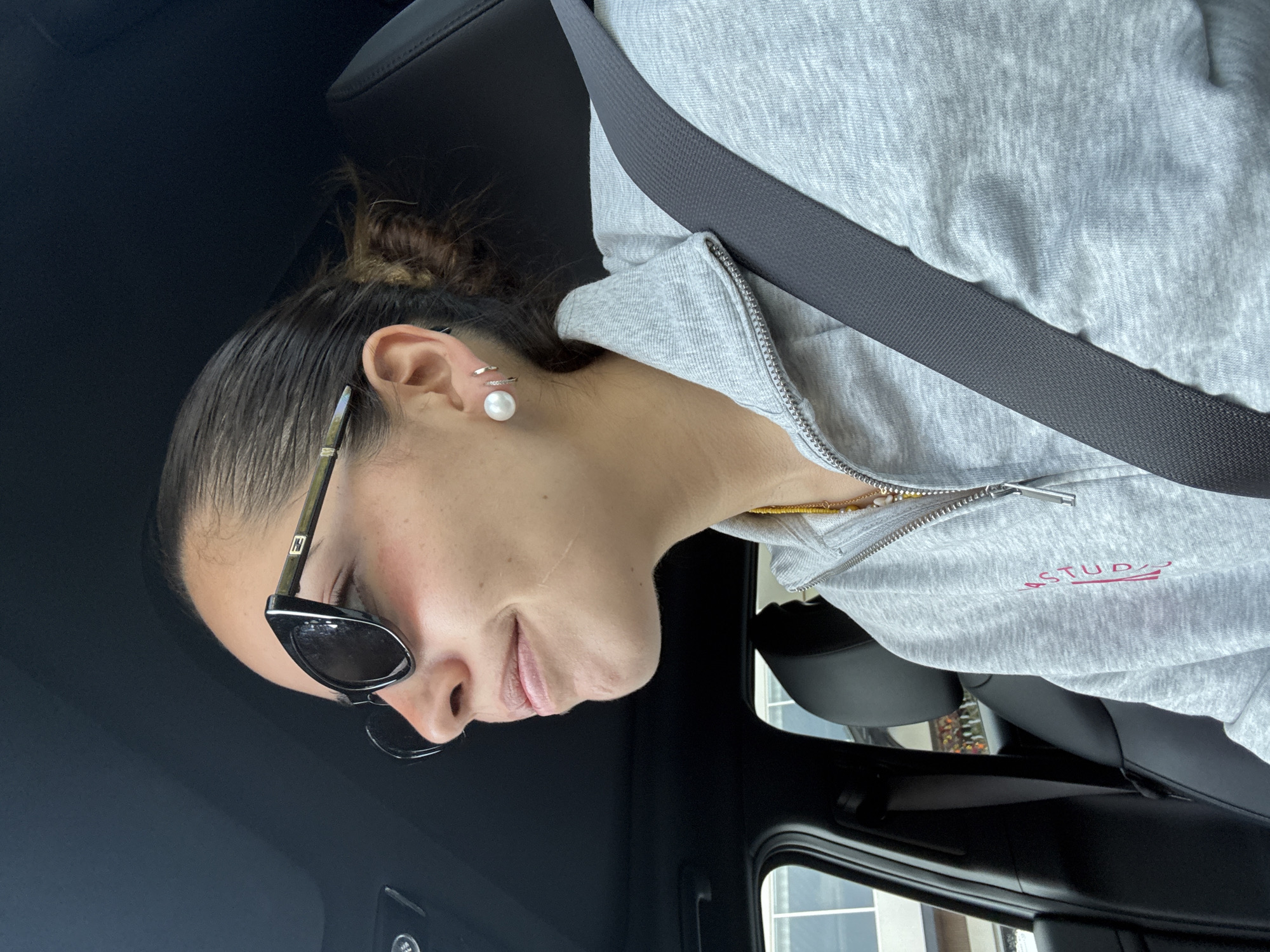 A woman wearing sunglasses, a gray zip-up hoodie with a "STUDIO" graphic, and a pearl earring is seated in a car. She has her seatbelt on and her hair is pulled back in a bun. The car appears to be parked, and a building is visible through the window. It is not possible to determine the specific location within the city of Puebla, Mexico from this image.