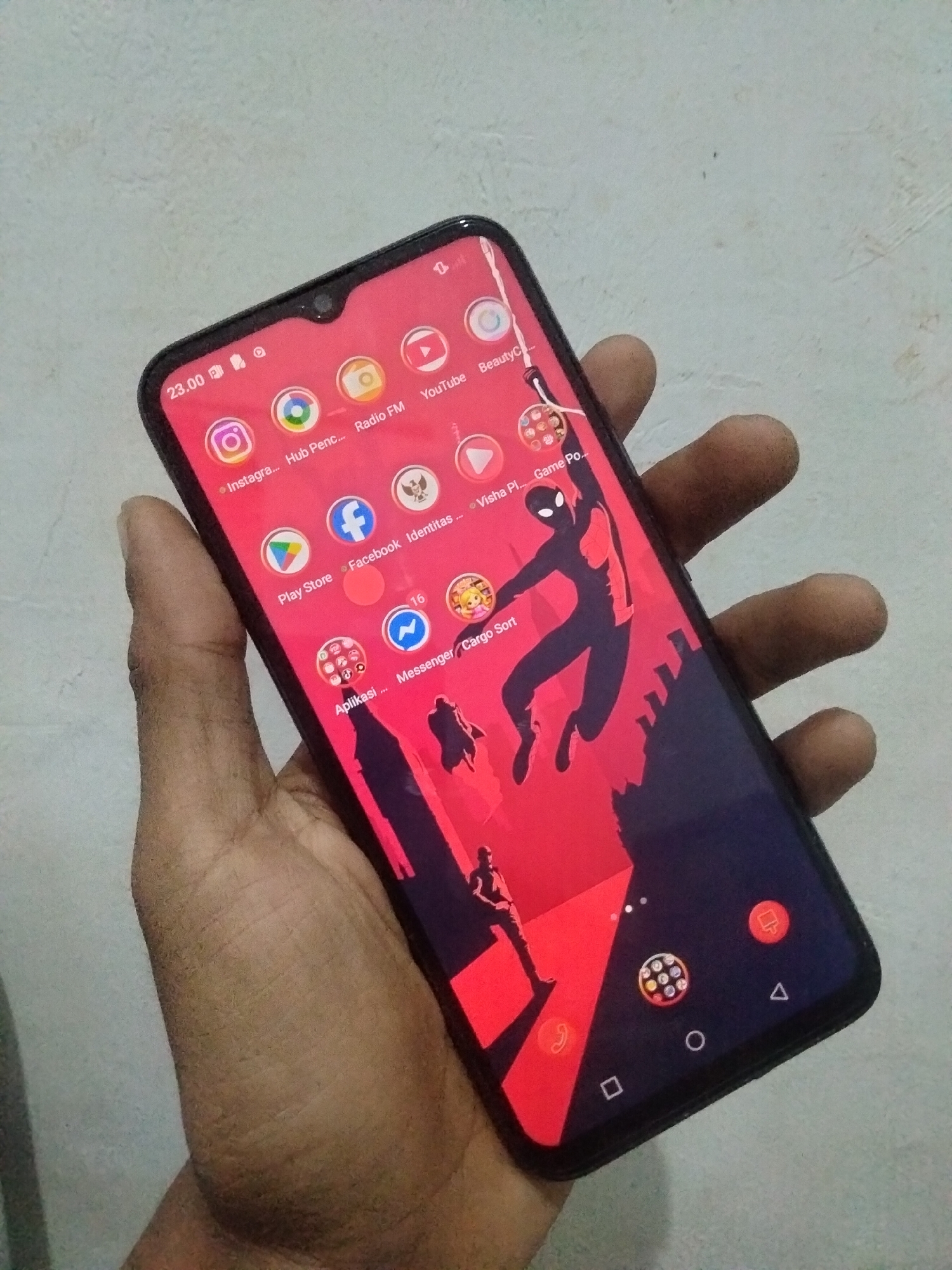 A hand holds a black smartphone with a red wallpaper displaying a silhouette of Spider-Man. The phone's screen shows various app icons, including Instagram, Facebook, YouTube, and Play Store. A clock at the top left displays "23.00". The background is a plain, light-colored surface. The image appears to have been taken in Antirogo, Indonesia, though no specific landmarks or city details are visible in the frame.