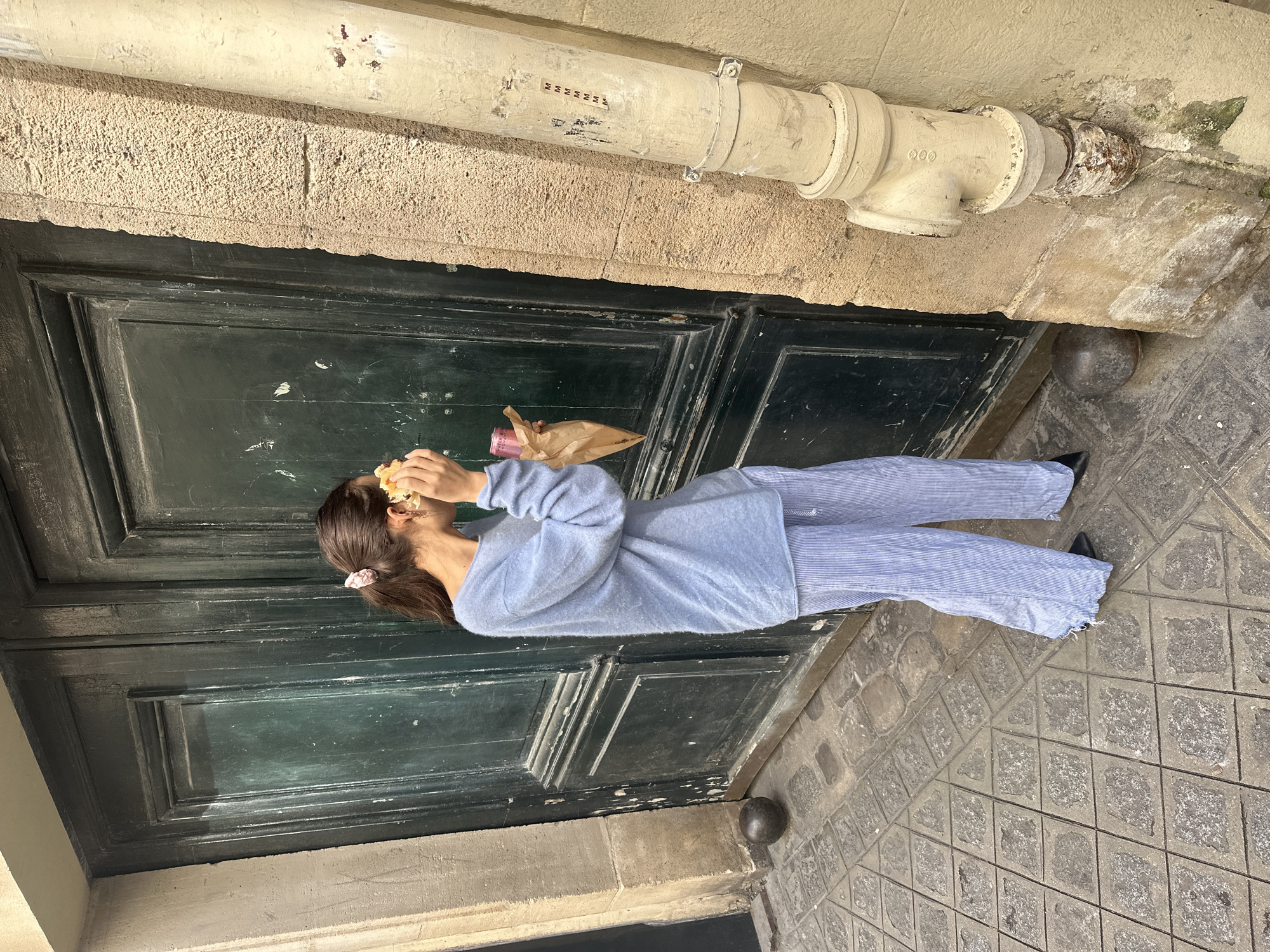 The image shows a person standing in front of a weathered, dark green, double door. The person, appearing to be female with brown hair tied back with a pink scrunchie, is wearing a light blue top and matching striped pants. The person is holding a sandwich or pastry in their hands, seemingly about to take a bite. A brown paper bag and a cylindrical item (possibly a drink container) are also being held by the person.
The door is flanked by aged stone walls. Above the door is a pipe affixed to the wall, showing signs of wear and oxidation. The ground is paved with patterned cobblestones.
Given the architectural style, paving stones, and overall aesthetic, it is possible this scene is located in Paris, France.